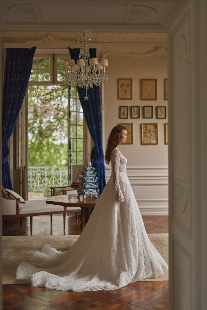 Woman standing in a room wearing a long white lace wedding dress with a train