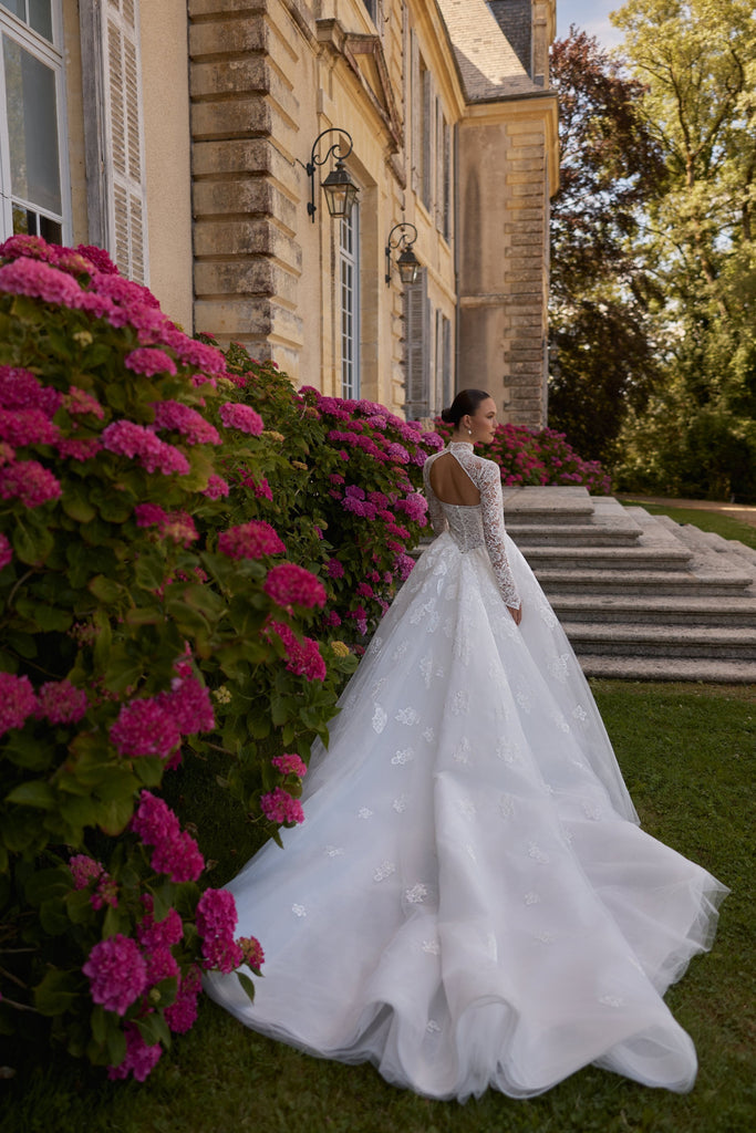 Bride in long sleeve lace wedding dress with train standing by pink hydrangea bushes and stone building
