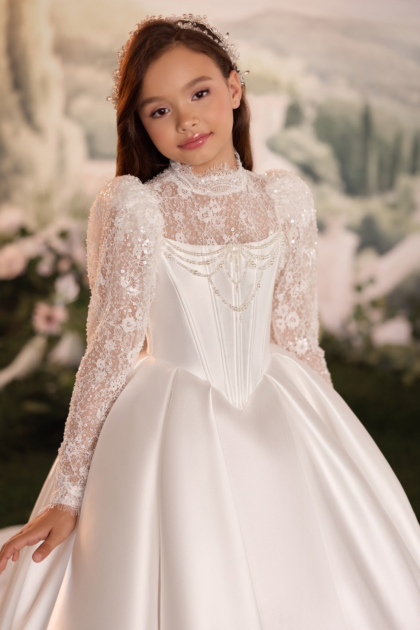 A young girl wearing an elegant white ball gown with long lace sleeves, a high mock neck, and a full satin skirt, posed against a soft, blurred scenic background.