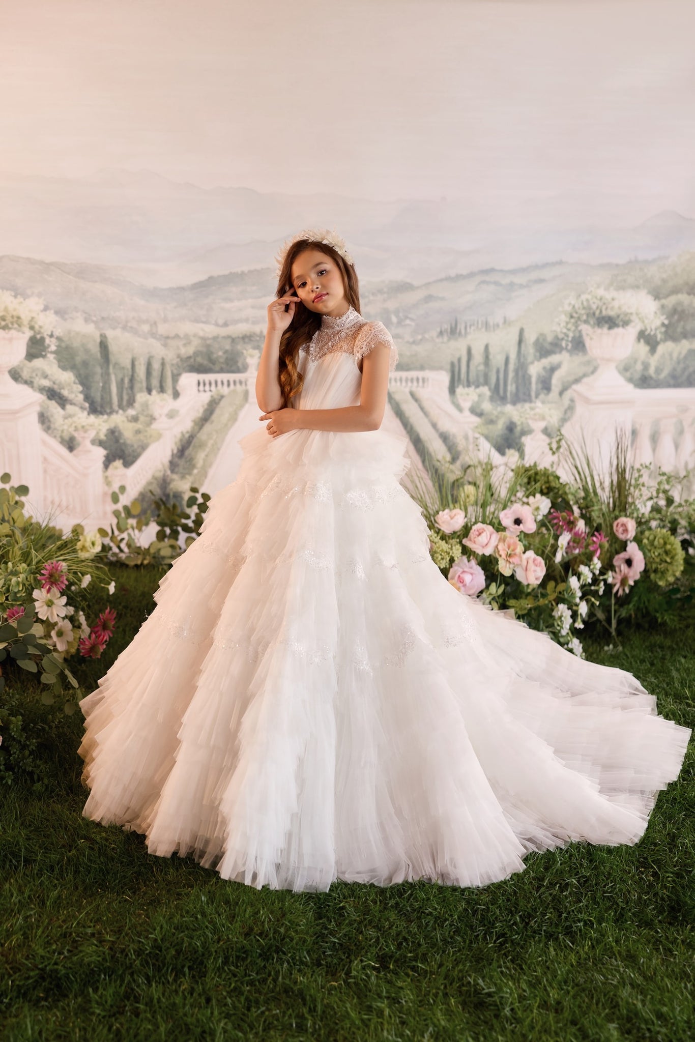 Girl wearing a white tiered tulle gown with lace details and a floral headpiece standing in a garden setting