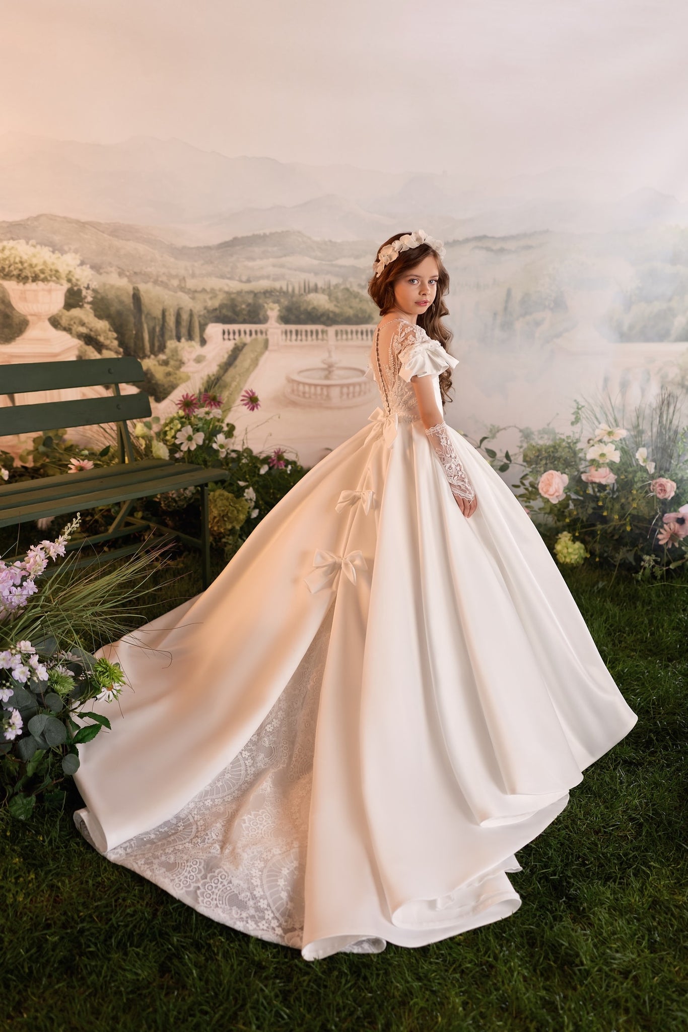 Girl wearing a white satin ball gown with lace details and a floral headband standing in a garden setting