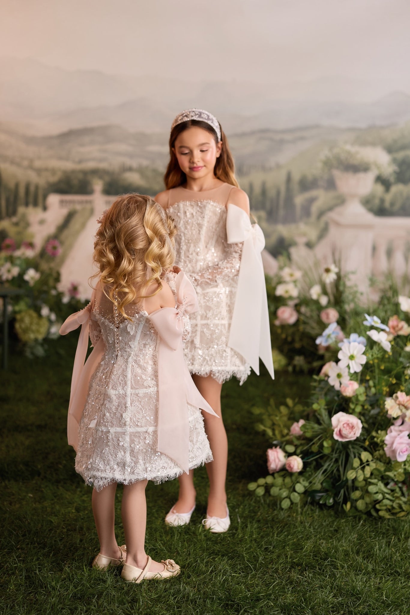 Two girls wearing matching embellished short dresses with bows standing in a garden setting