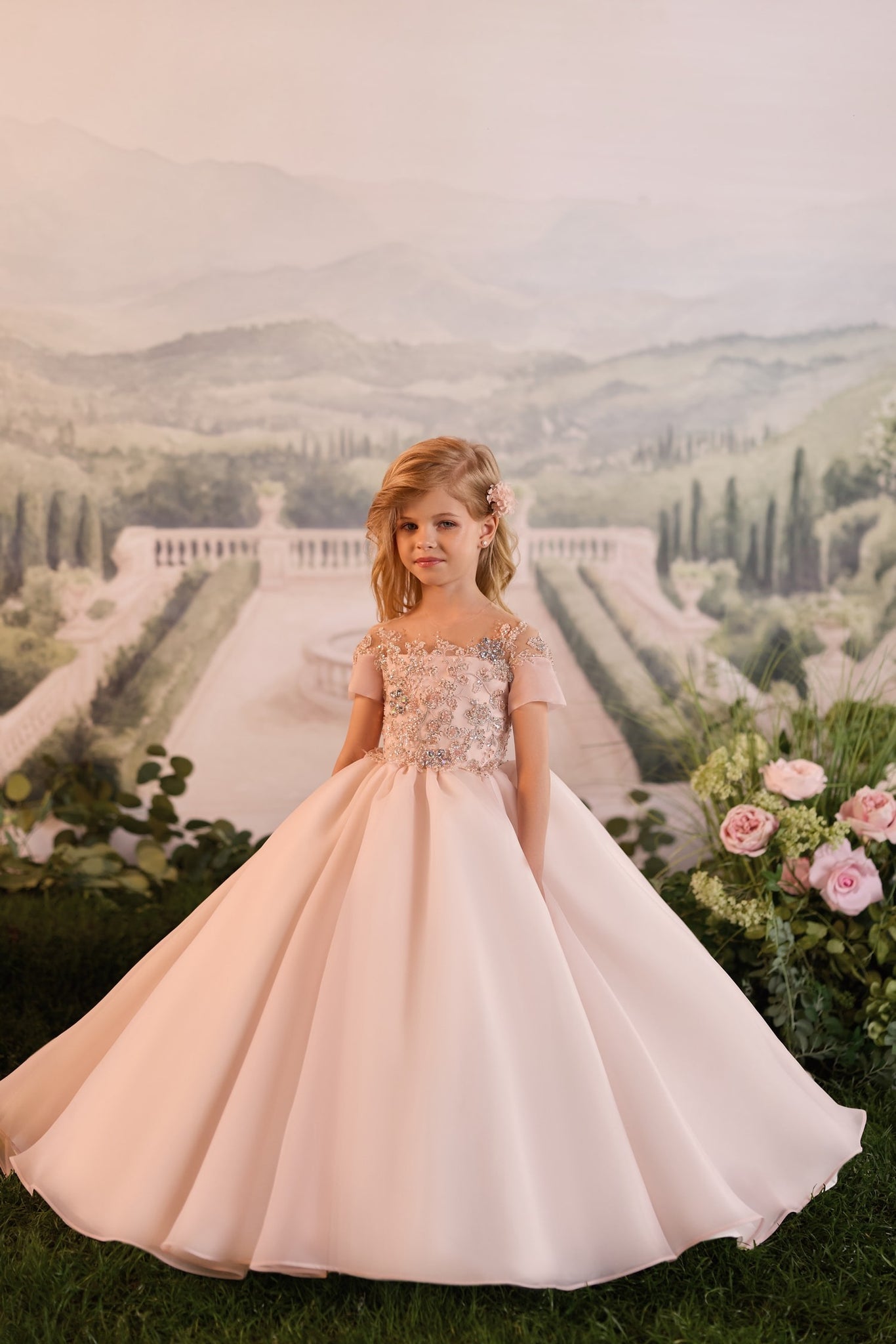 A young girl stands in a formal ball gown with a full skirt, photographed in front of a scenic garden backdrop featuring a long path, hills, and pink flowers. The dress is pale blush pink with a beaded embellished bodice and short sheer sleeves.
