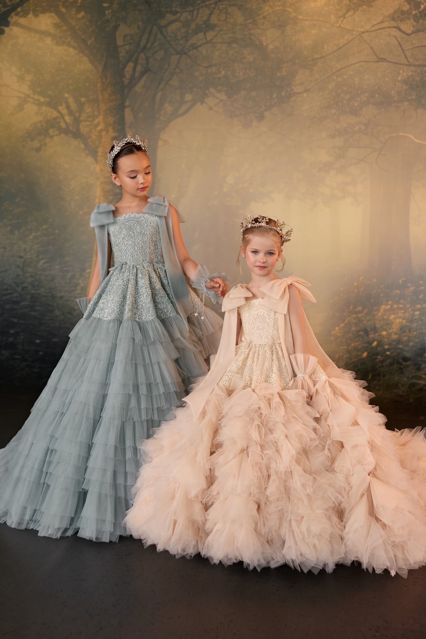 Two girls wearing floor length tulle gowns and crowns standing in front of a forest backdrop