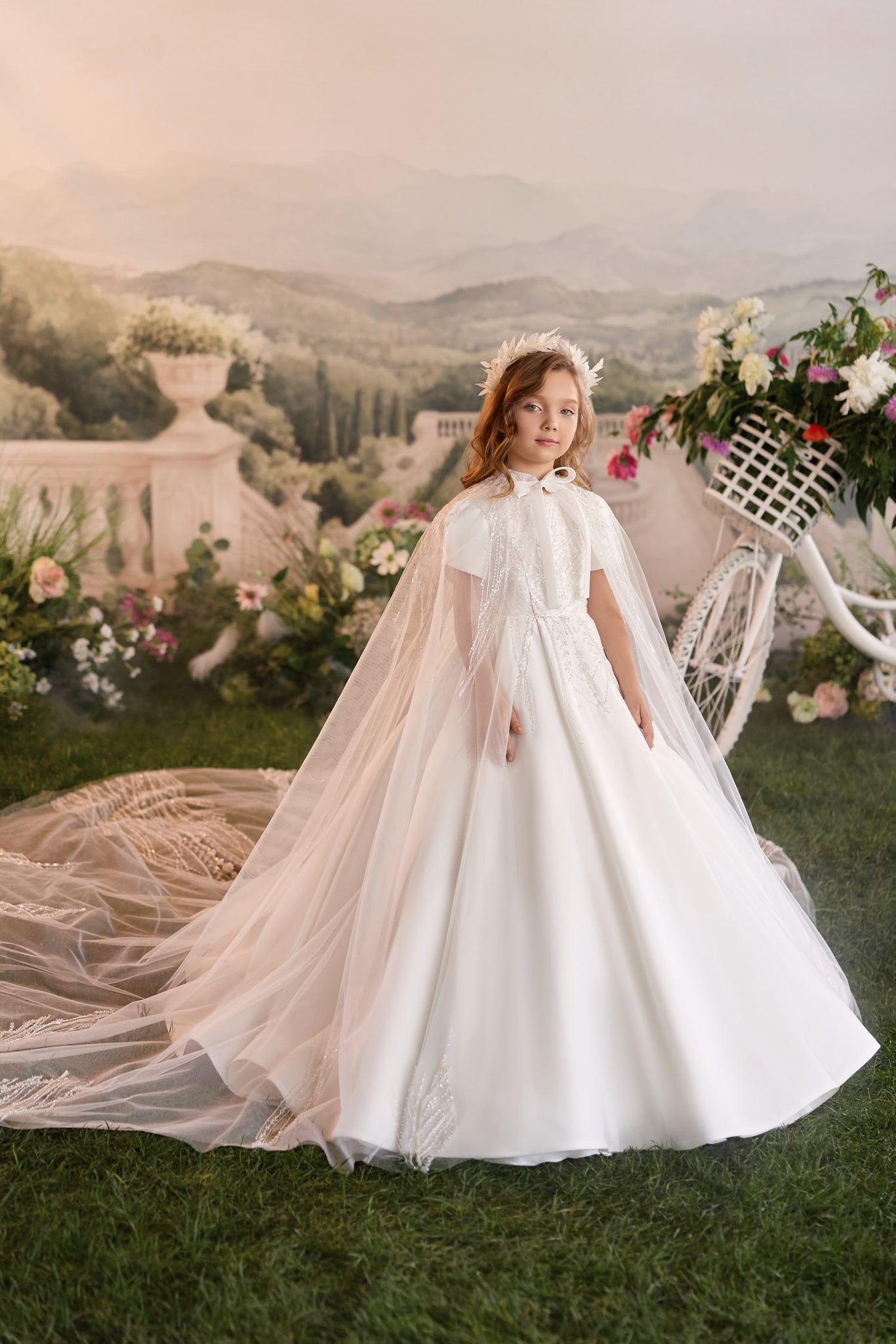 Girl wearing a white communion dress with a long cape and floral headpiece standing in a garden setting