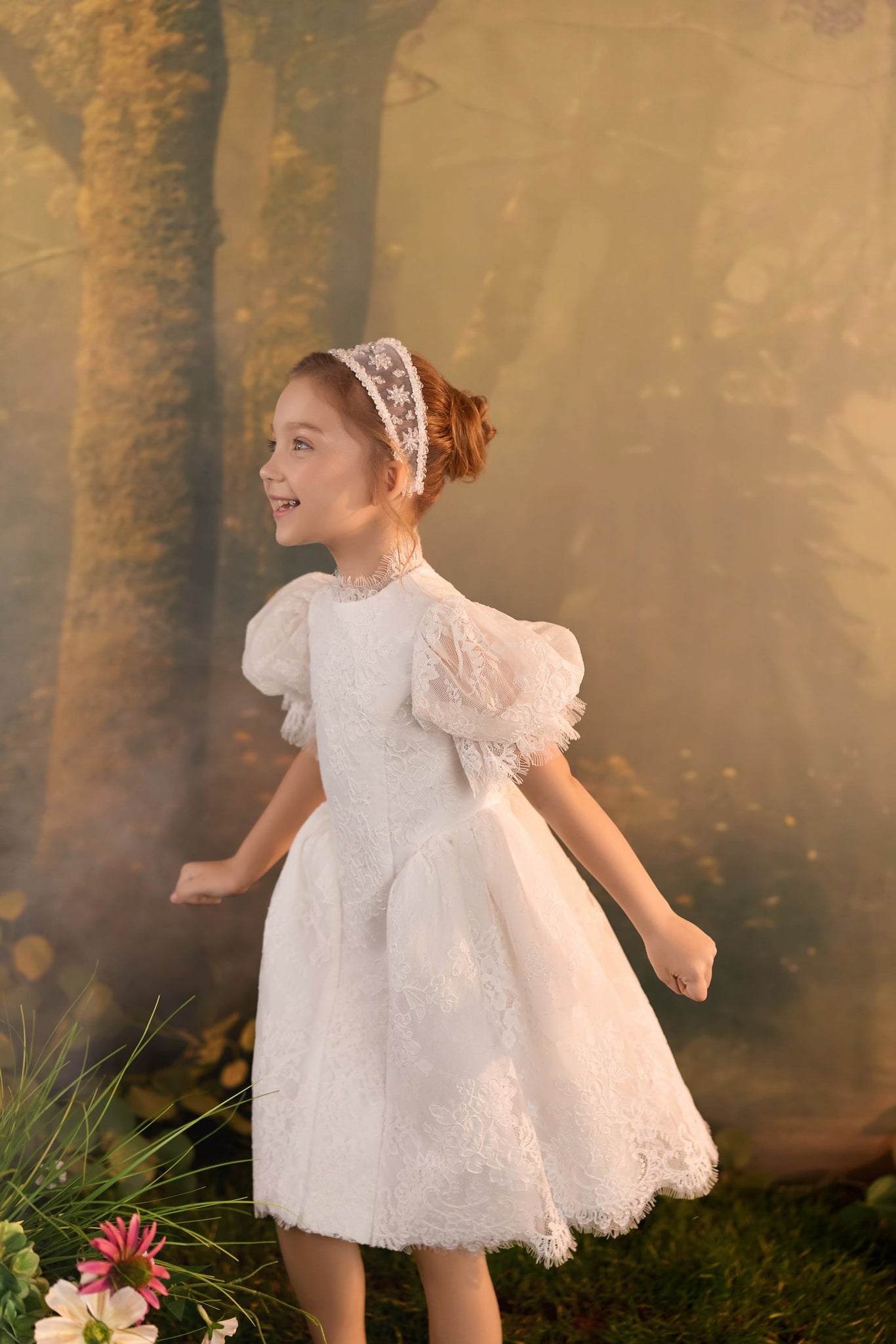 Girl wearing a white lace dress and a matching headband standing in front of a forest backdrop | Girls Formal Dress