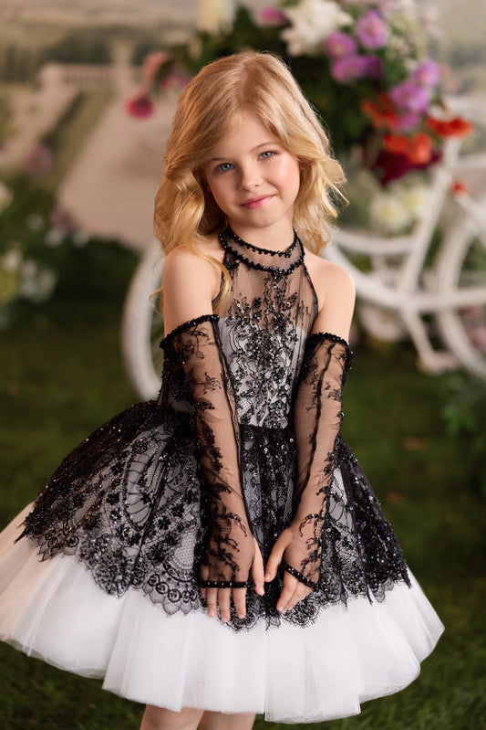 A young girl with blonde hair wearing a black and white lace and tulle formal dress, standing in front of a blurred garden background with flowers and a white chair.