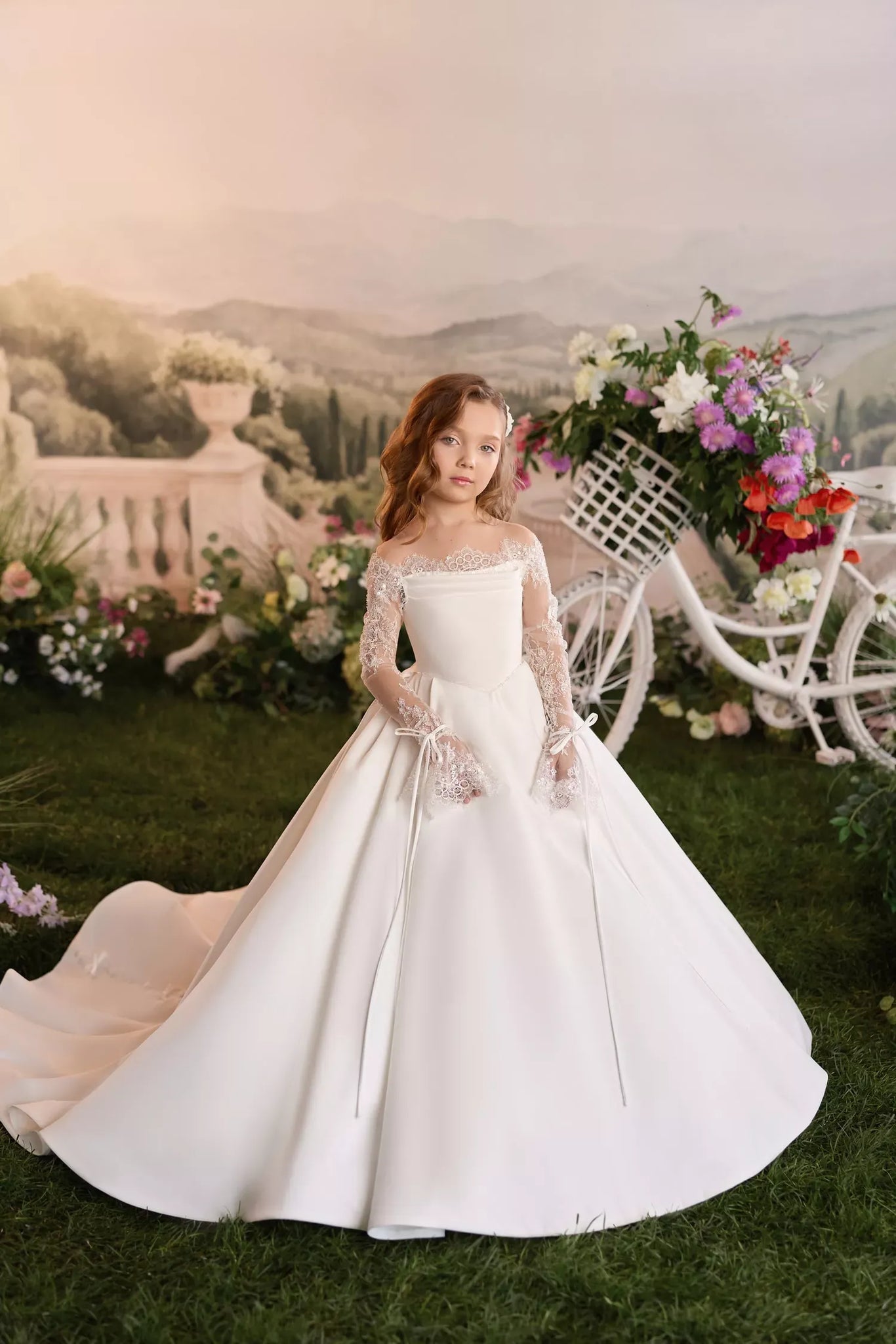 Girl wearing a white off the shoulder ball gown with lace long sleeves standing in a garden with a flower bicycle