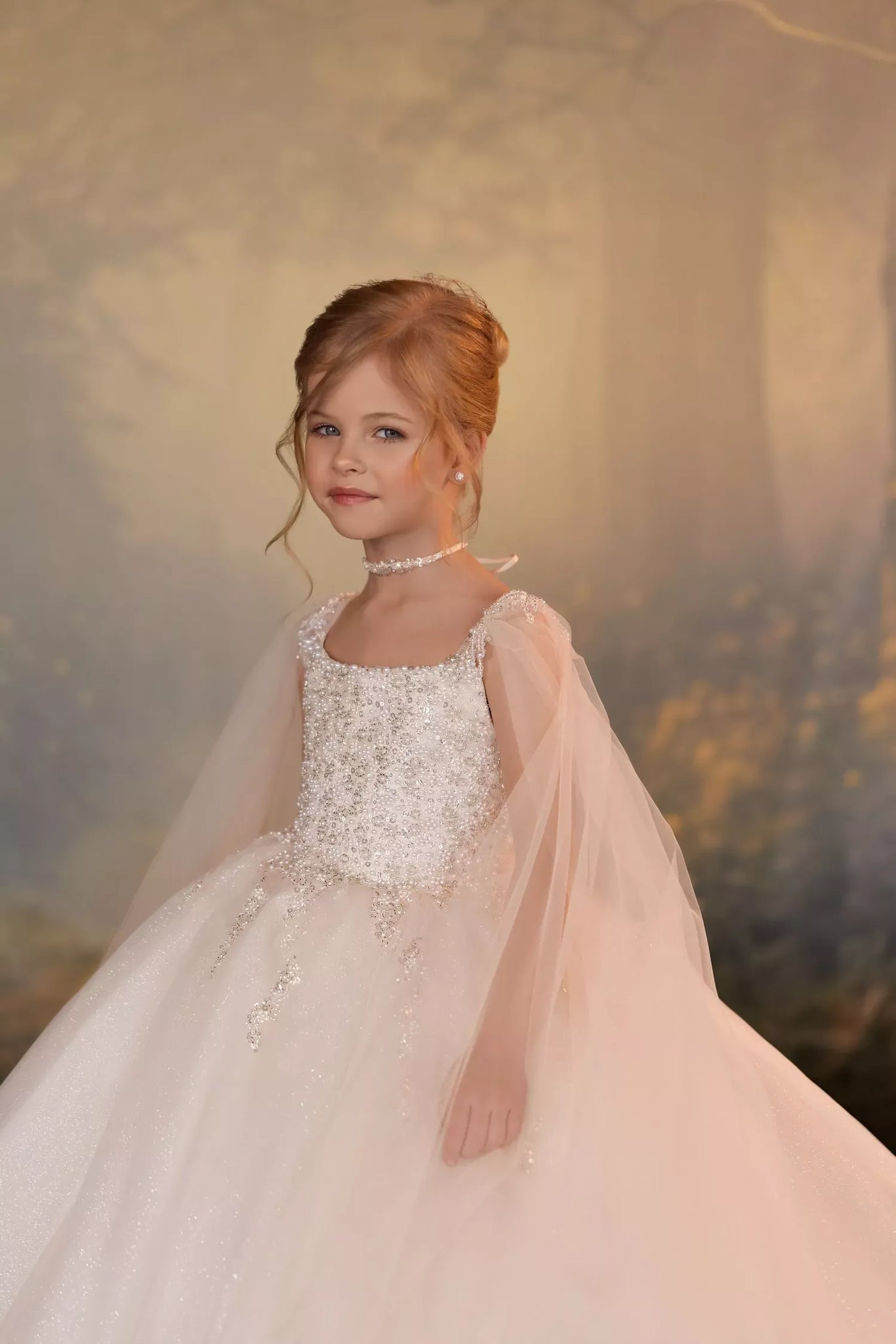 A young girl with styled auburn hair wears a formal white and pale pink ball gown with a sheer cape and floral lace bodice, posed against a soft, warm-toned forest backdrop.