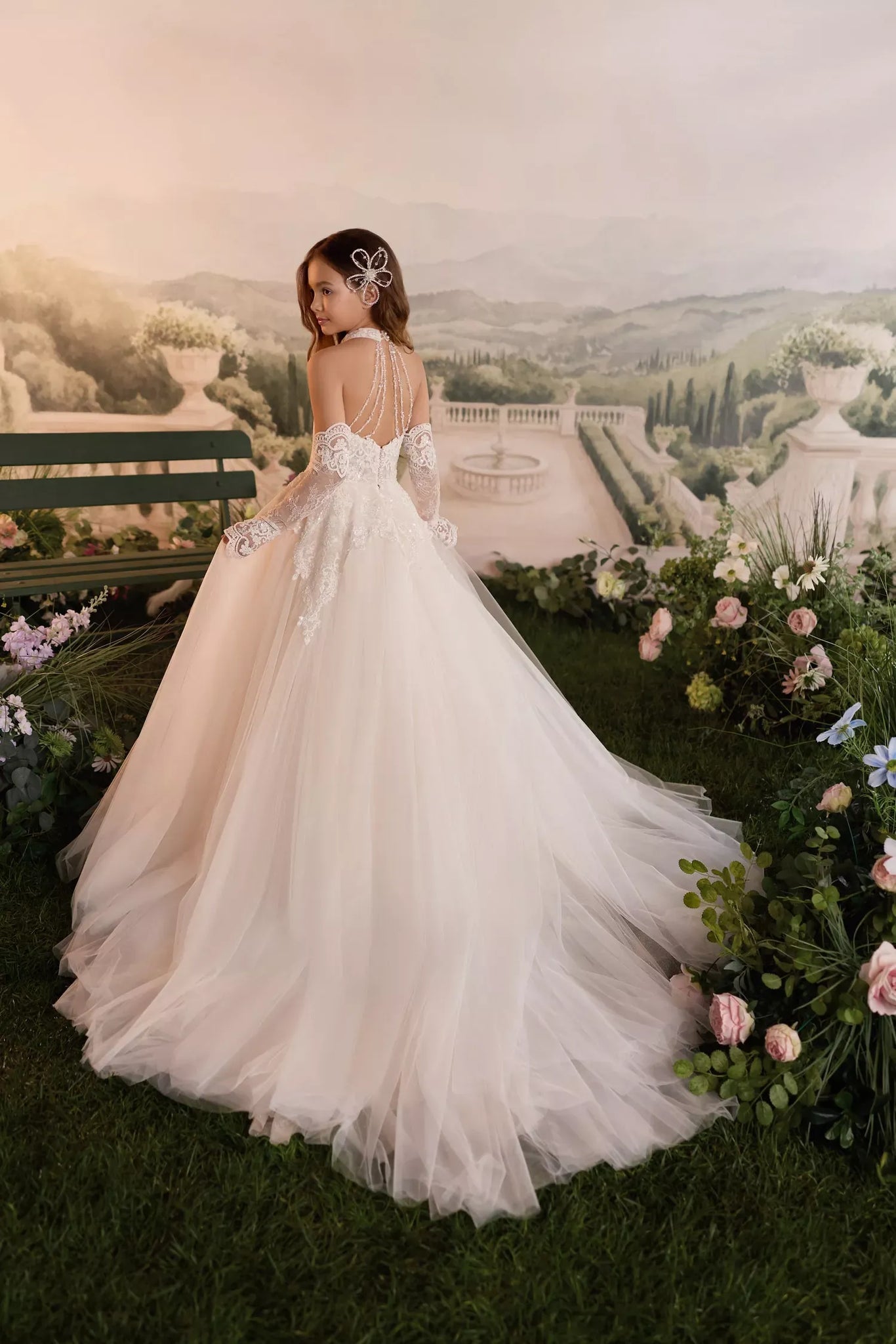 Girl wearing a white lace ball gown with a long tulle train and floral hair accessory in a garden setting | Girls Formal Dress