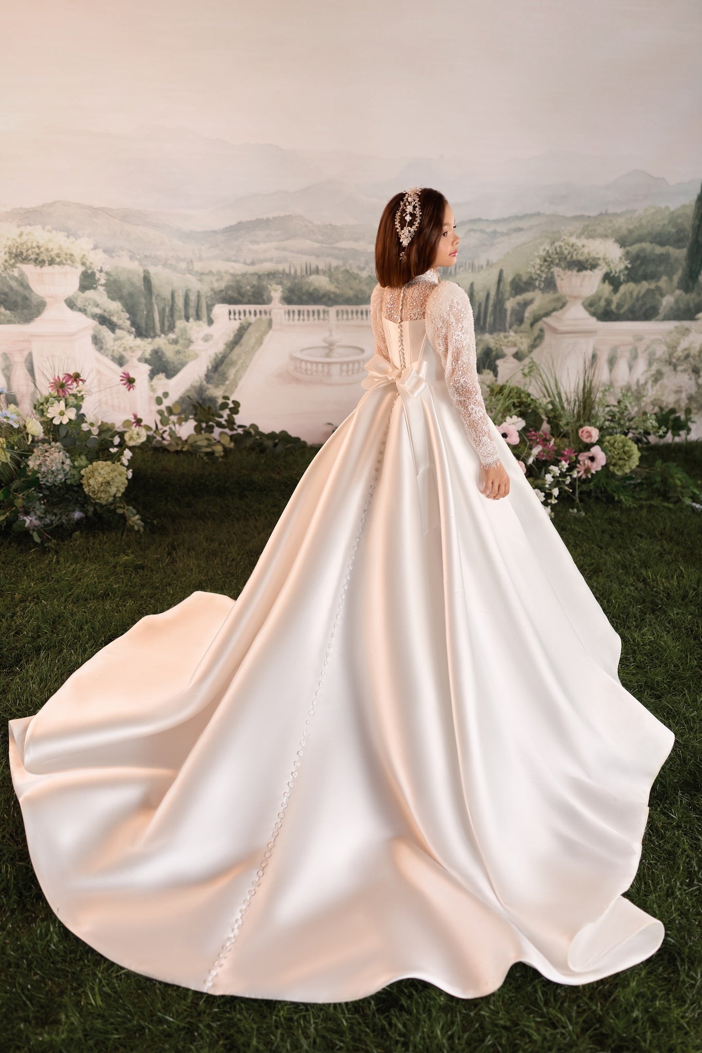 A girl wearing a voluminous ivory wedding gown with long lace sleeves and a sheer back, photographed from behind as she stands on grass in front of a scenic garden backdrop.