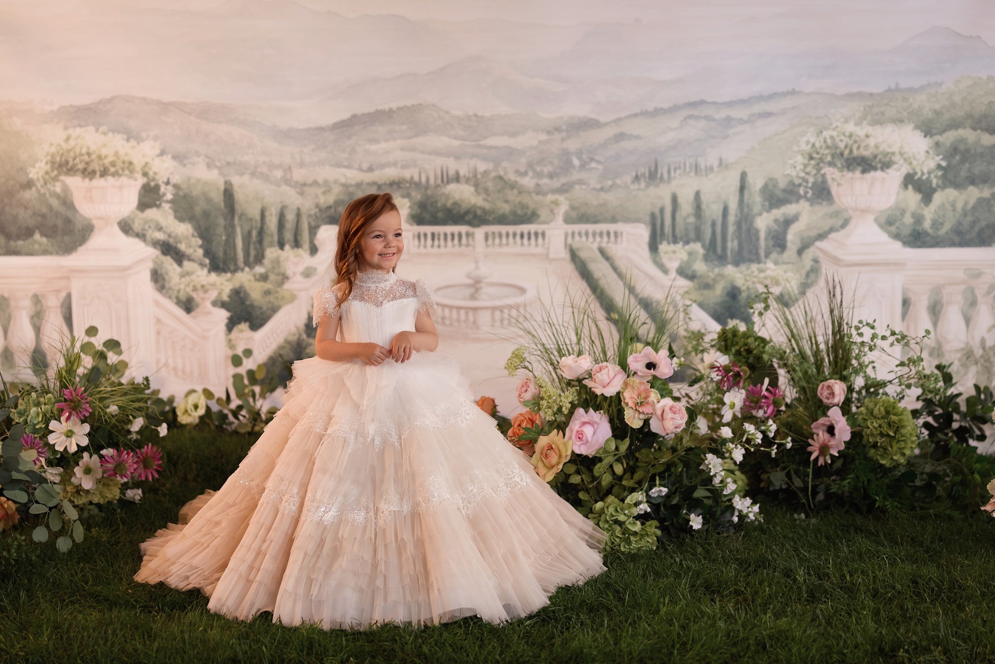 A young girl with red hair wearing a voluminous champagne-colored tulle ball gown stands in front of a scenic backdrop depicting a classical villa and lush green hills, flanked by large arrangements of pink, peach, and white flowers.