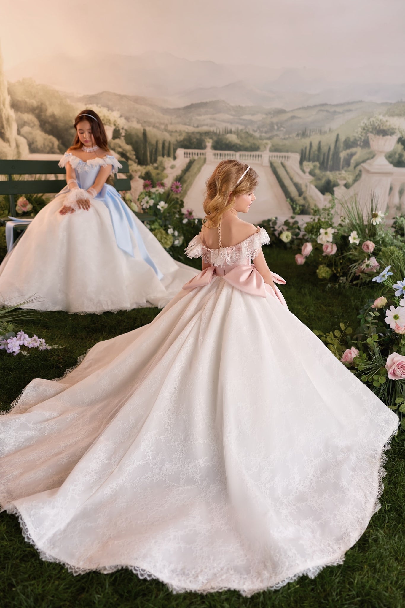 Two girls wearing elaborate ball gowns with lace details and long trains in a garden setting