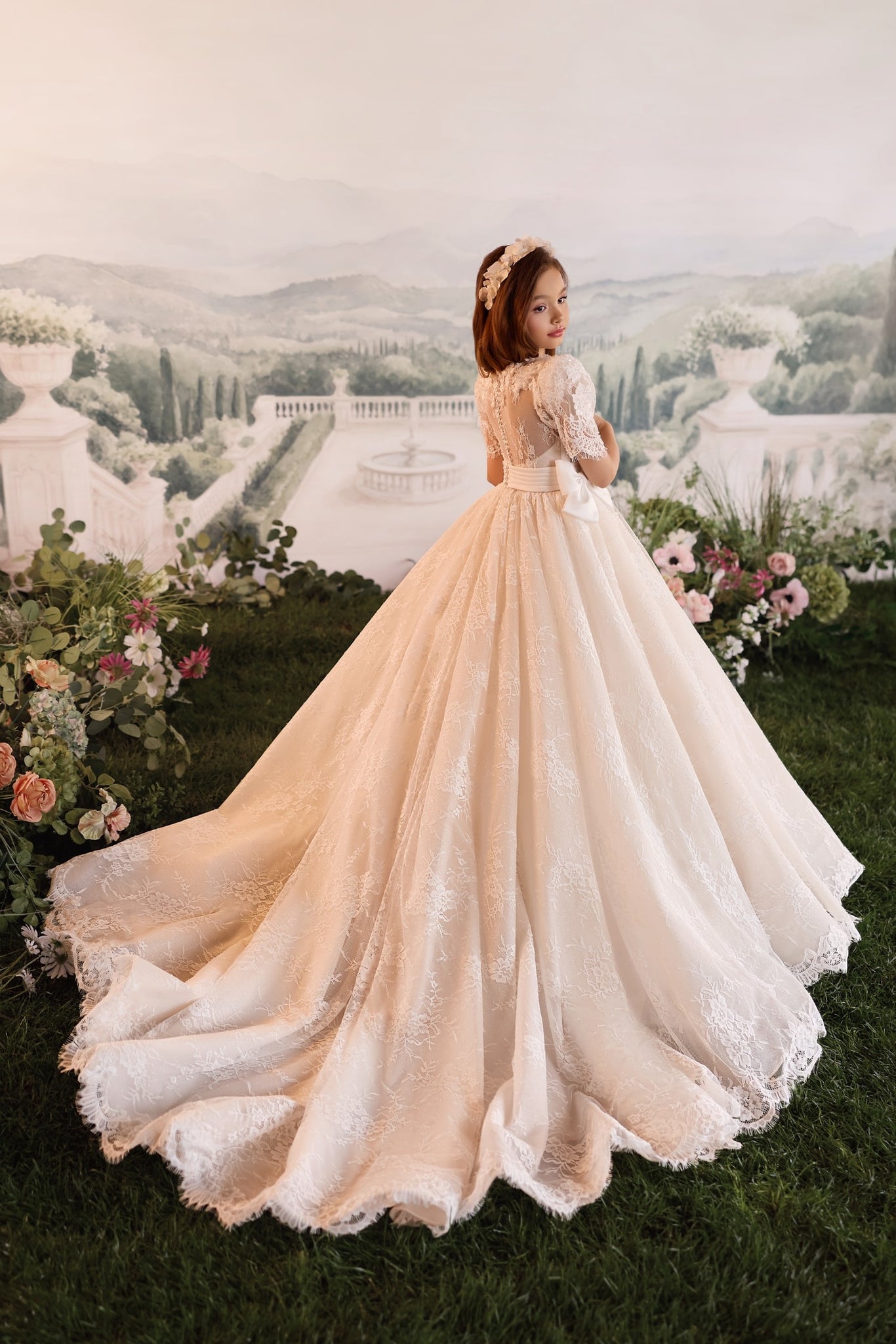 Girl wearing a white lace ball gown with a train and floral headband standing in a garden setting