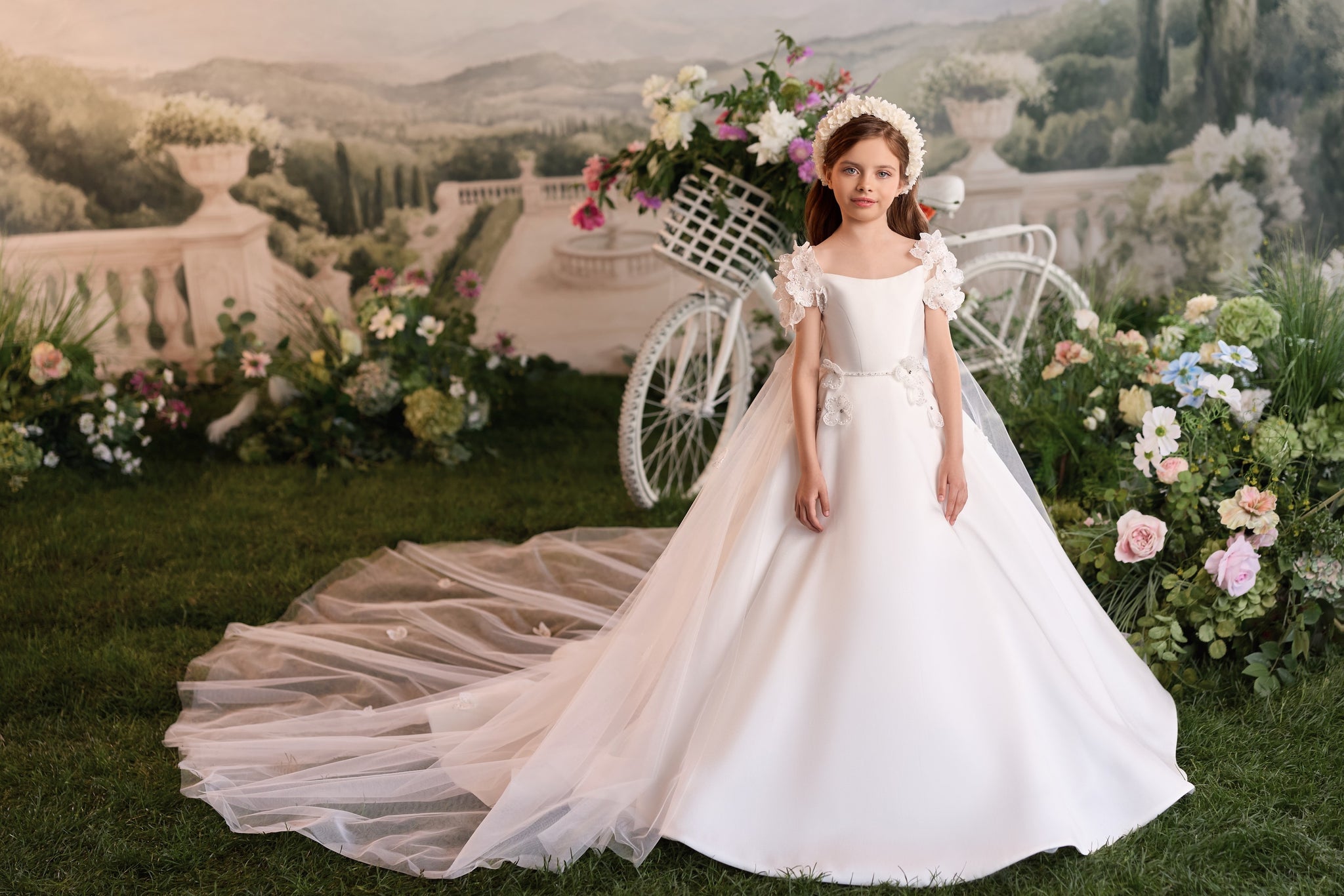 Girl wearing a white floor length dress with a long train and floral headpiece standing in front of a white bicycle