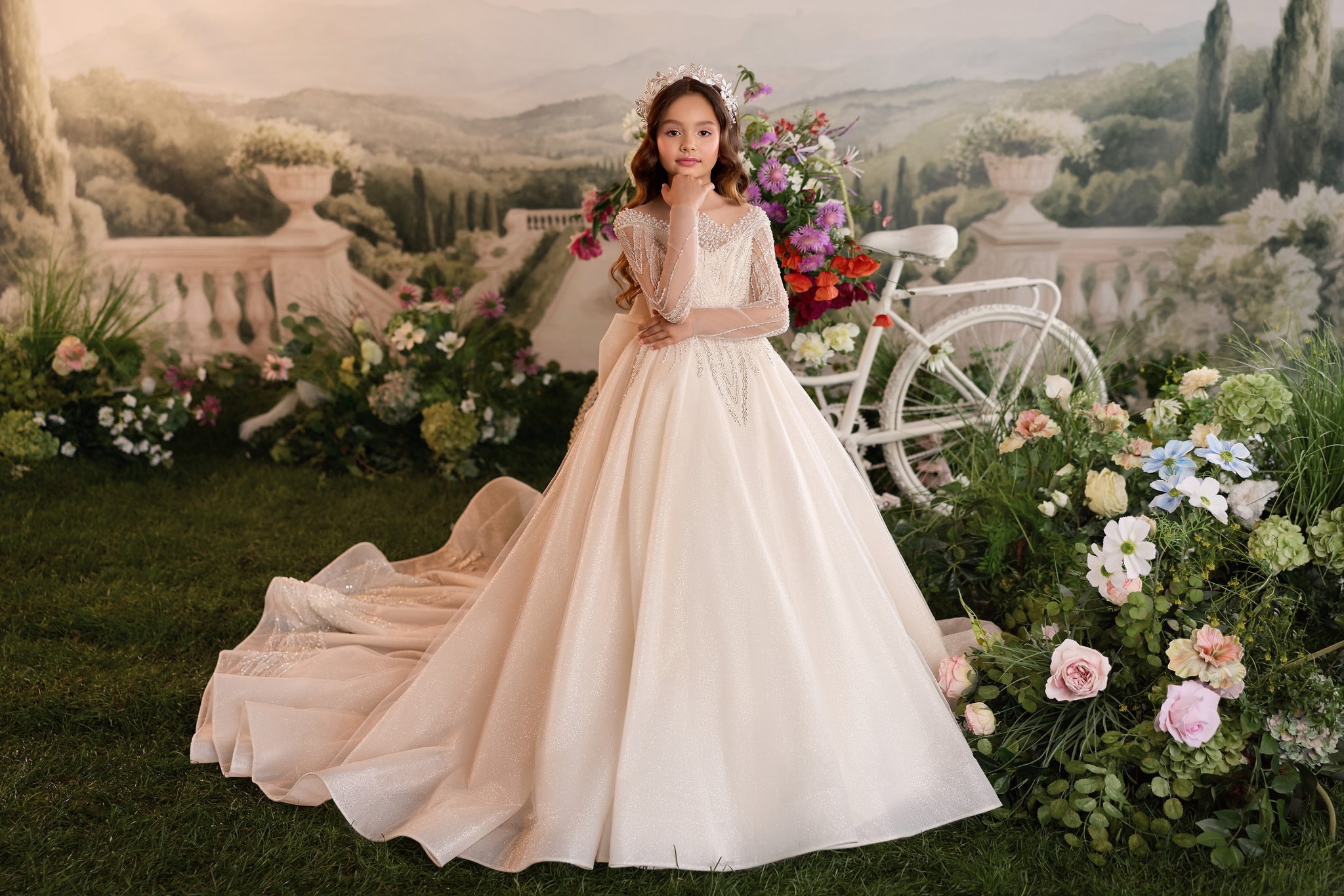 Girl wearing a long cream ball gown with a tiara standing in a garden setting with a white bicycle