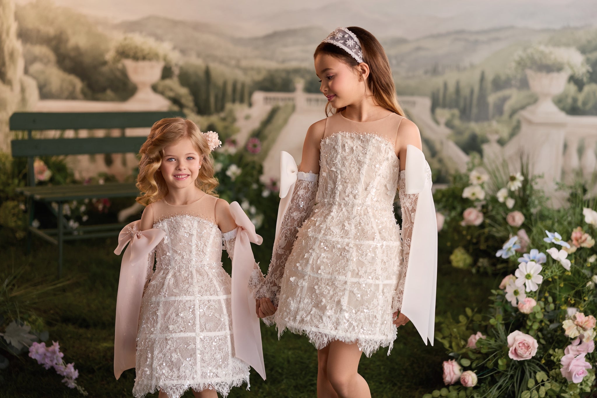 Two young girls wearing matching white lace dresses with bows and long sleeves standing in a garden setting