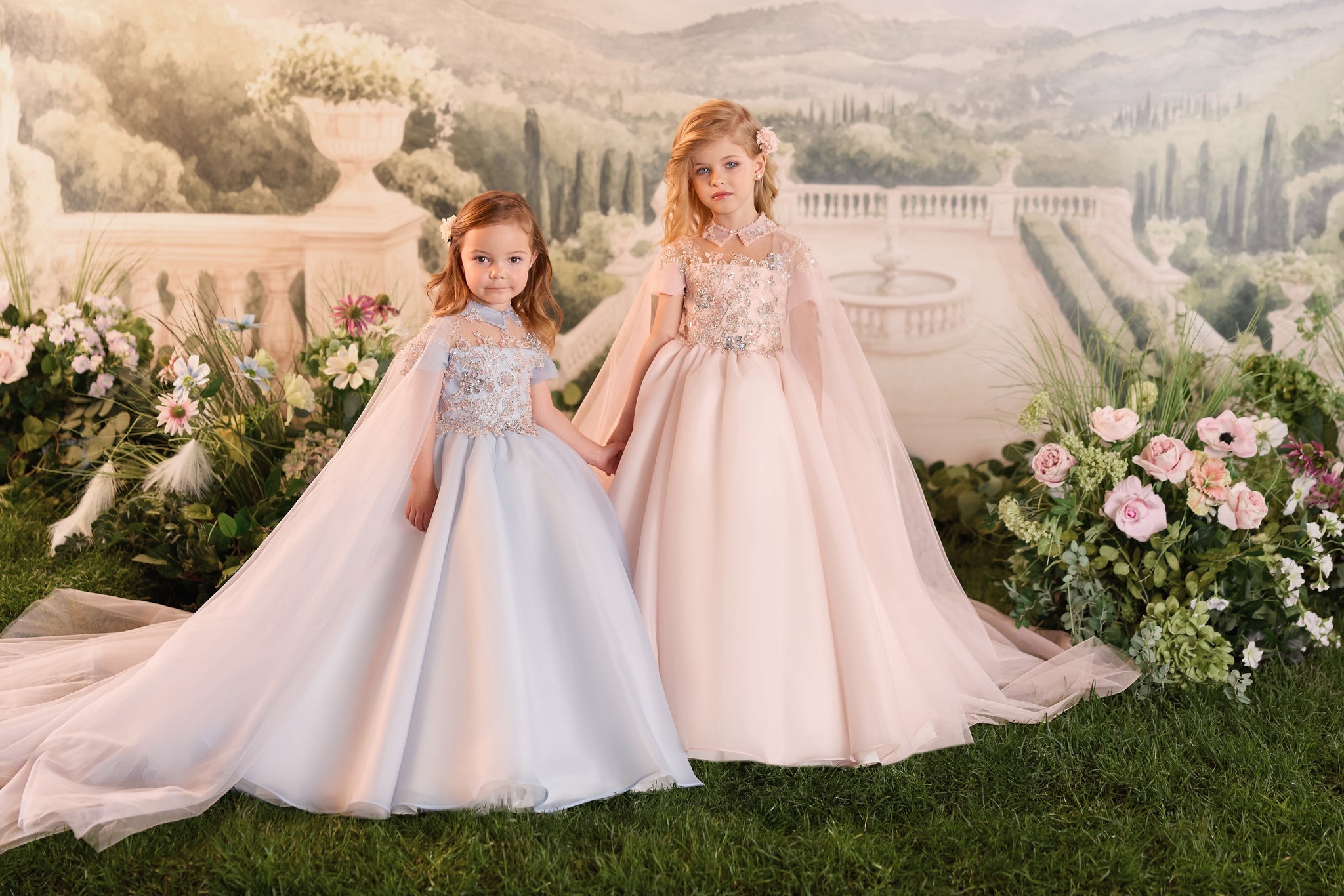 Two young girls wearing embellished floor length ball gowns with capes standing in a garden setting