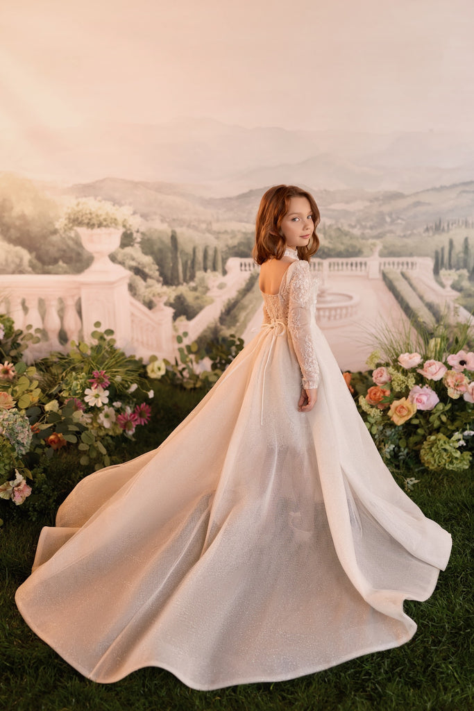 Girl wearing a convertible lace dress with a shimmering overskirt creating a floor-length gown and long train in a garden setting | Girls Formal Dress