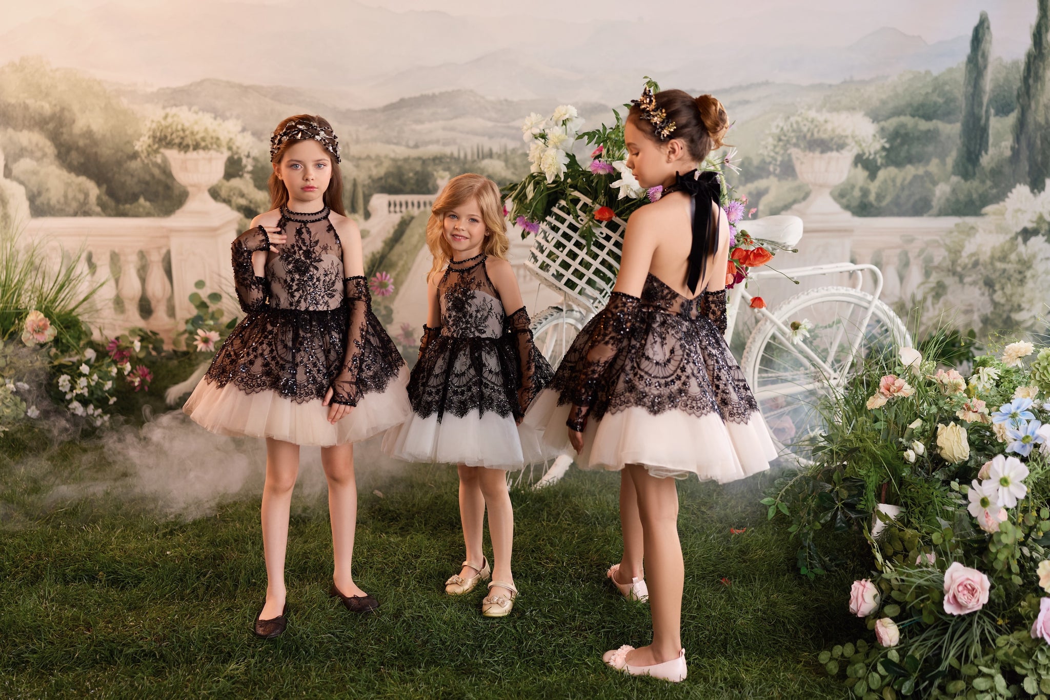 Three girls wearing black lace and beige tulle dresses standing in a garden setting with a white bicycle | Girls Formal Dress