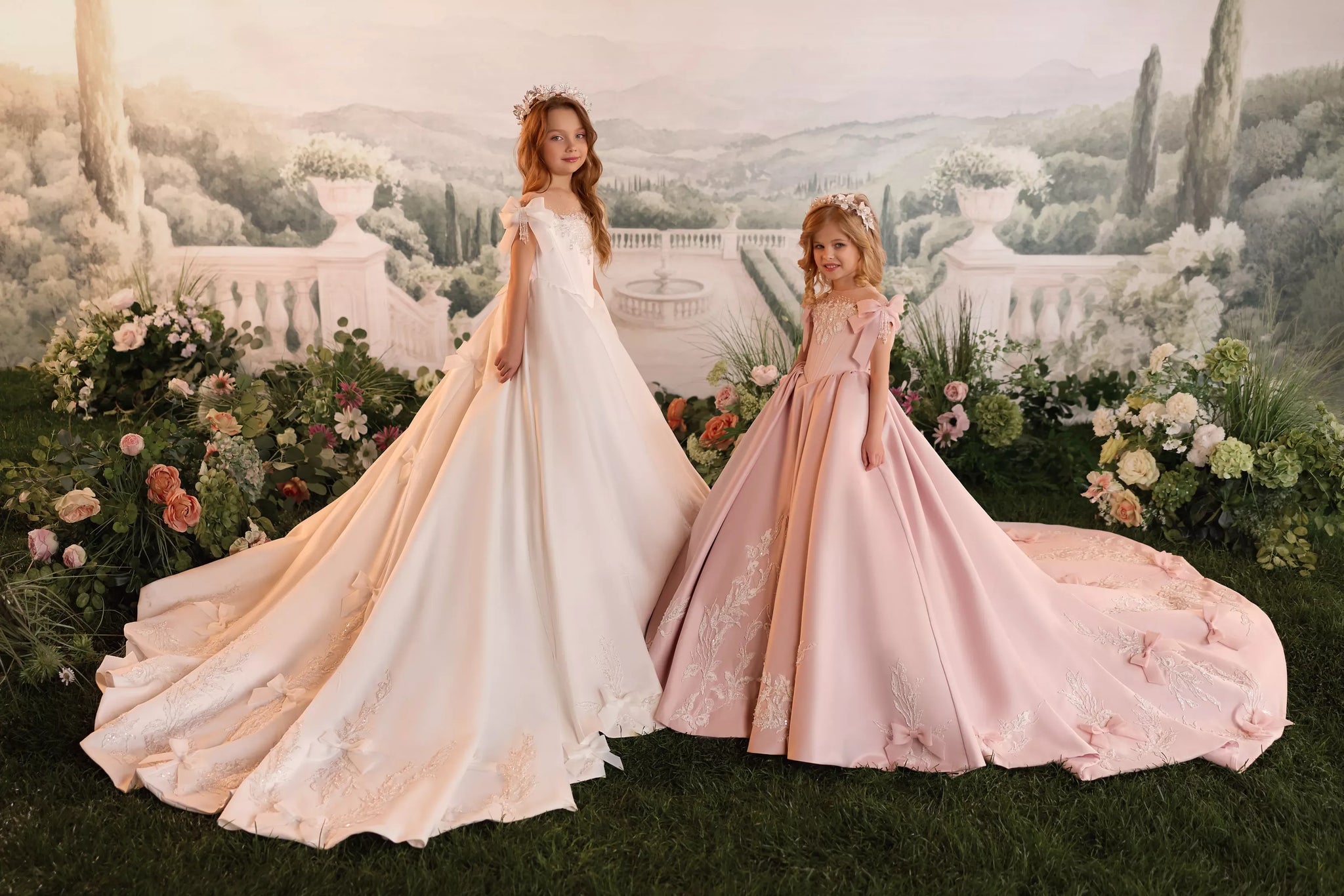 Two girls wearing white and pink ball gowns with floral crowns standing in a garden setting