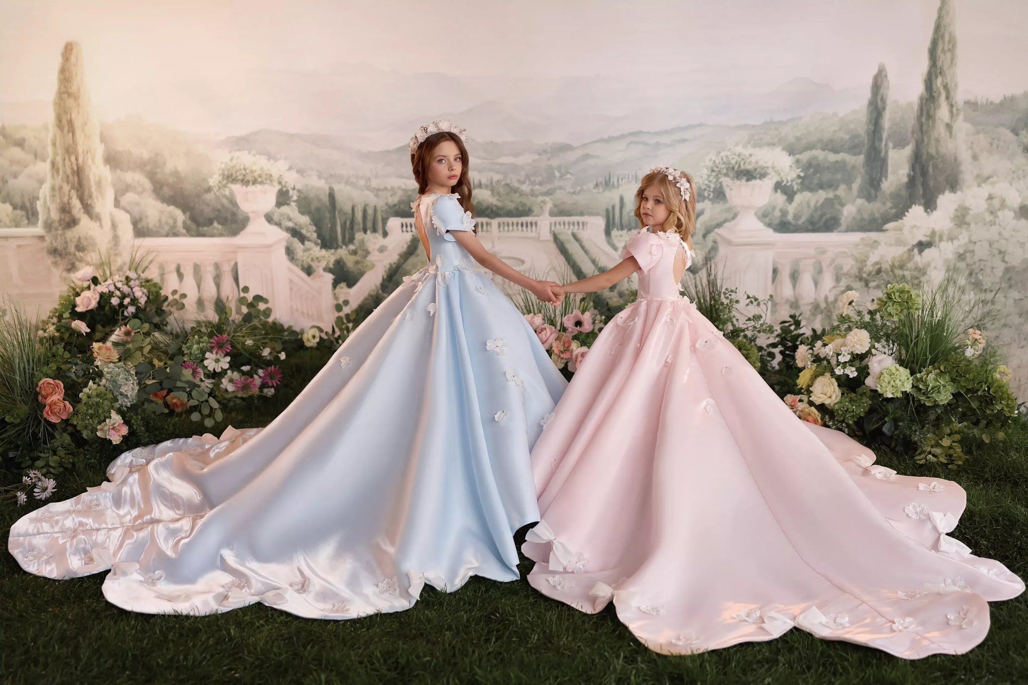 Two girls wearing long blue and pink ball gowns with floral accents standing in a garden setting