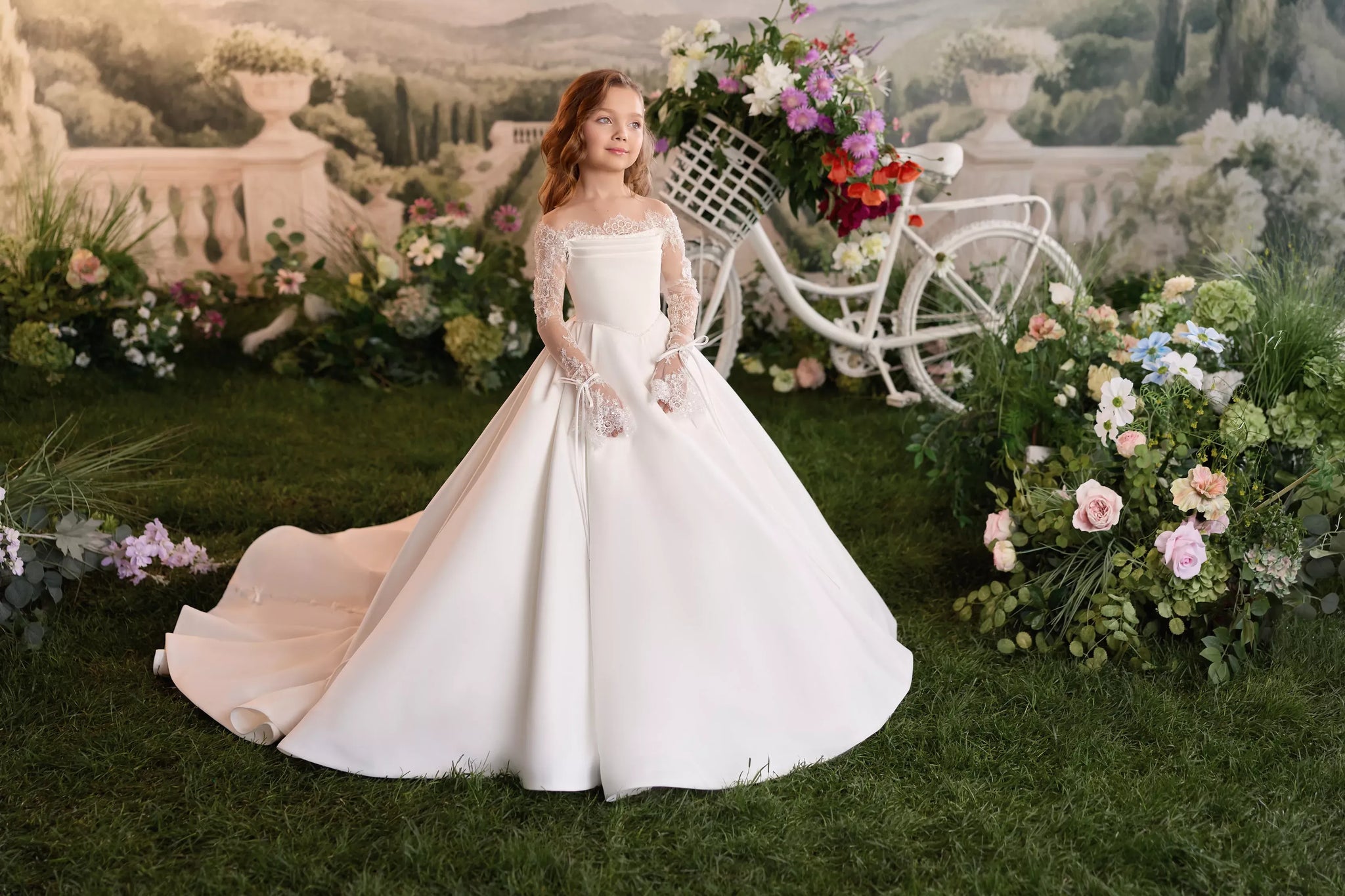 Girl wearing a white off-shoulder ball gown with lace sleeves standing in a garden with a decorative bicycle
