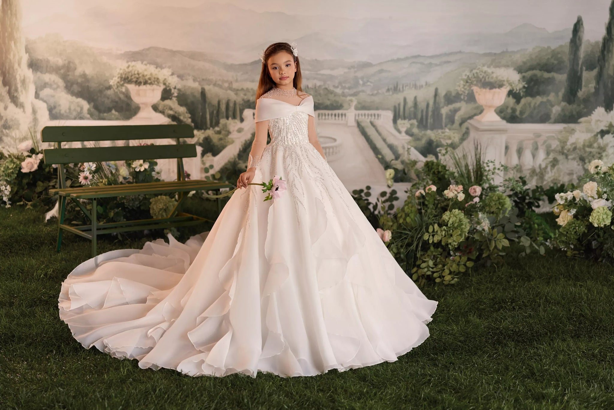 Girl wearing a white ball gown with a long train and beaded bodice holding a flower in a garden setting