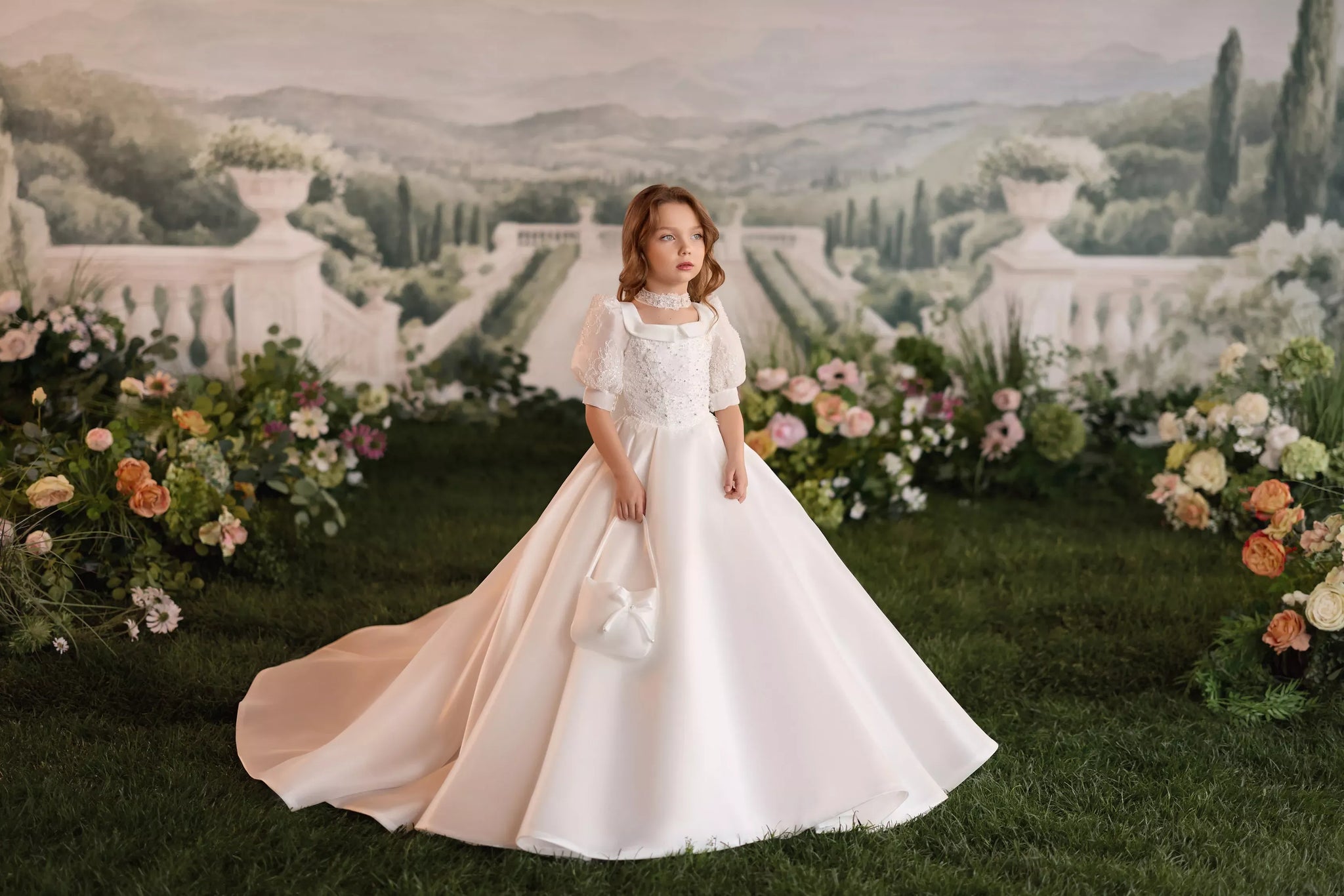 Girl wearing a white ball gown with puff sleeves and a matching handbag standing in a garden | Girls Formal Dress