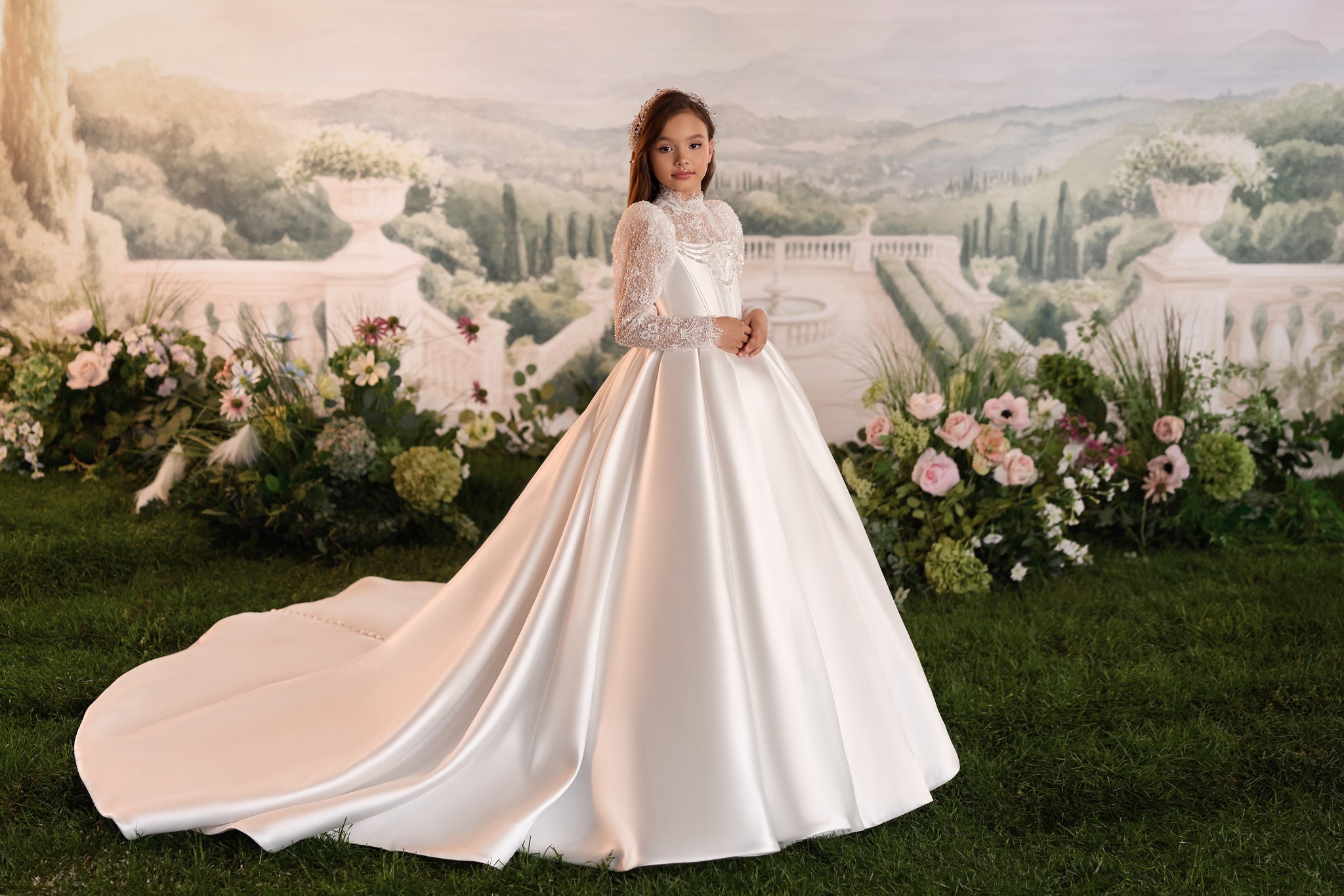A young girl wearing a white satin ball gown with lace sleeves stands on a grassy lawn in front of a scenic garden backdrop with flowers and a classical villa mural.
