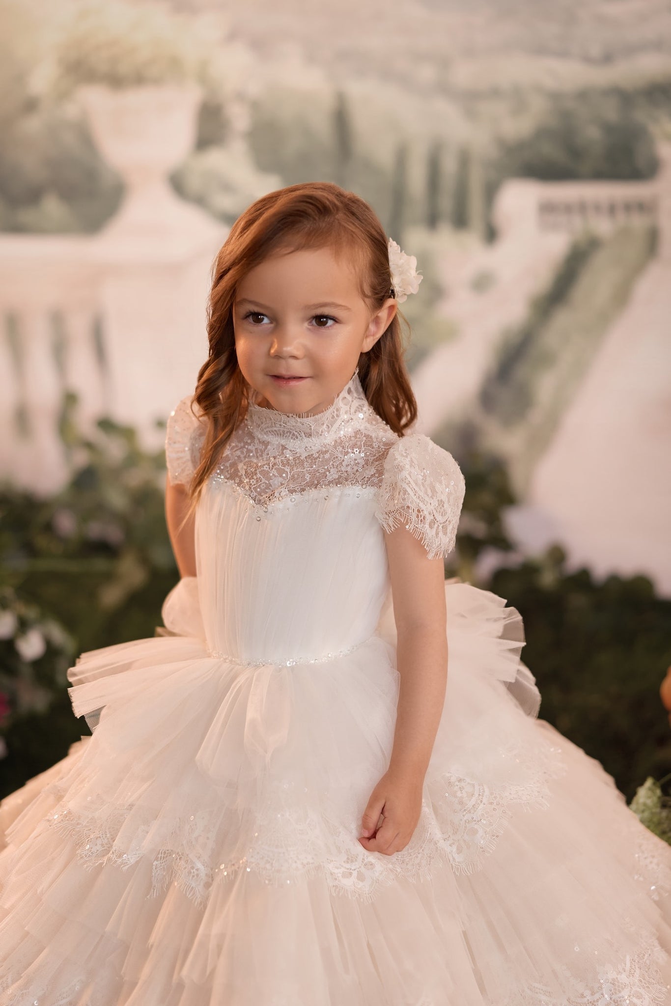A young girl with red hair wearing a white first communion dress with lace detailing and a tulle skirt, standing in front of a soft-focus garden backdrop.