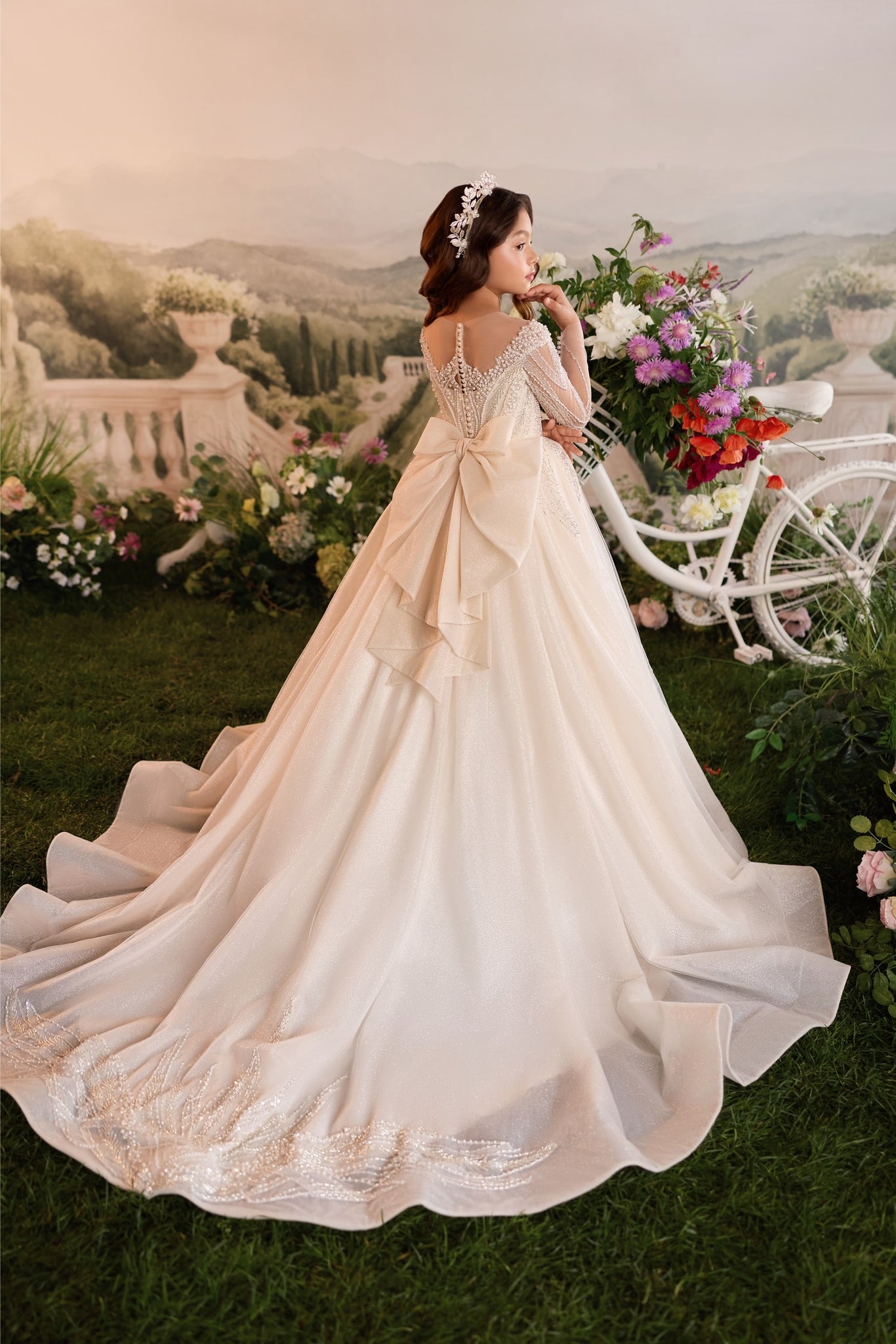 Girl wearing a long cream ball gown with a large bow and beaded details standing by a decorative bicycle