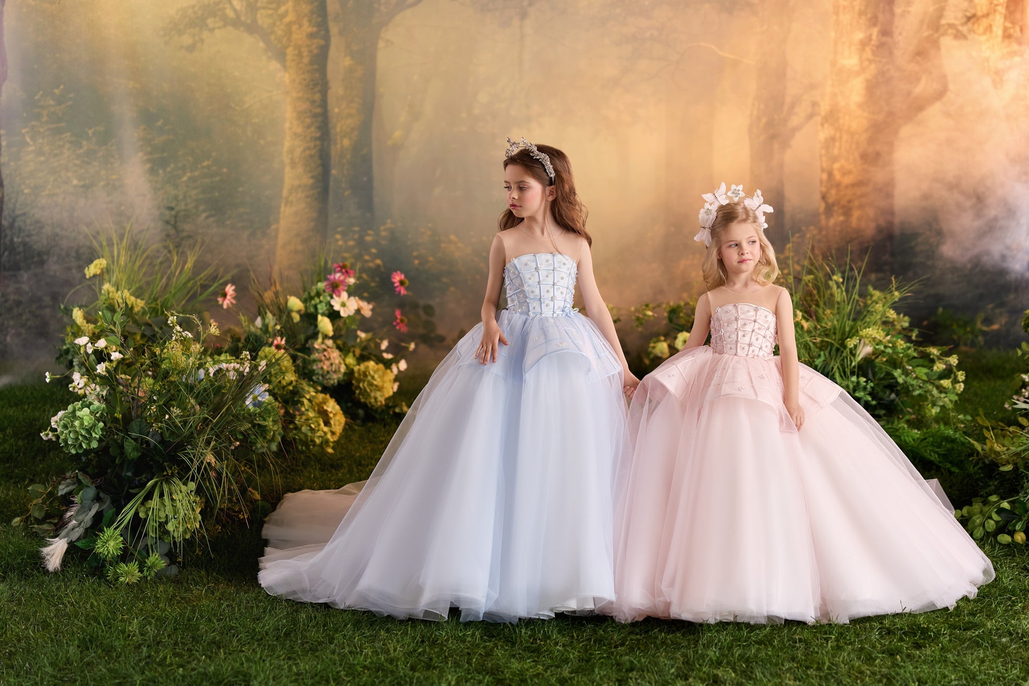 Two girls wearing pastel blue and pink ball gowns with floral headpieces standing in a forest setting