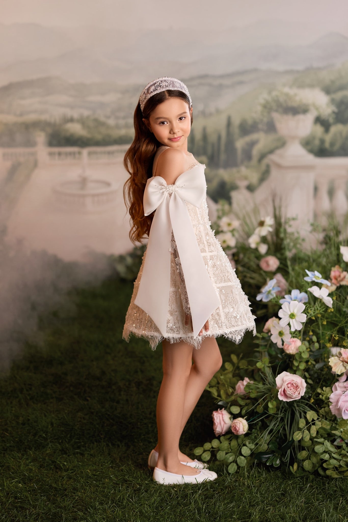 Girl wearing a white lace dress with a large bow and a beaded headband standing in a garden setting