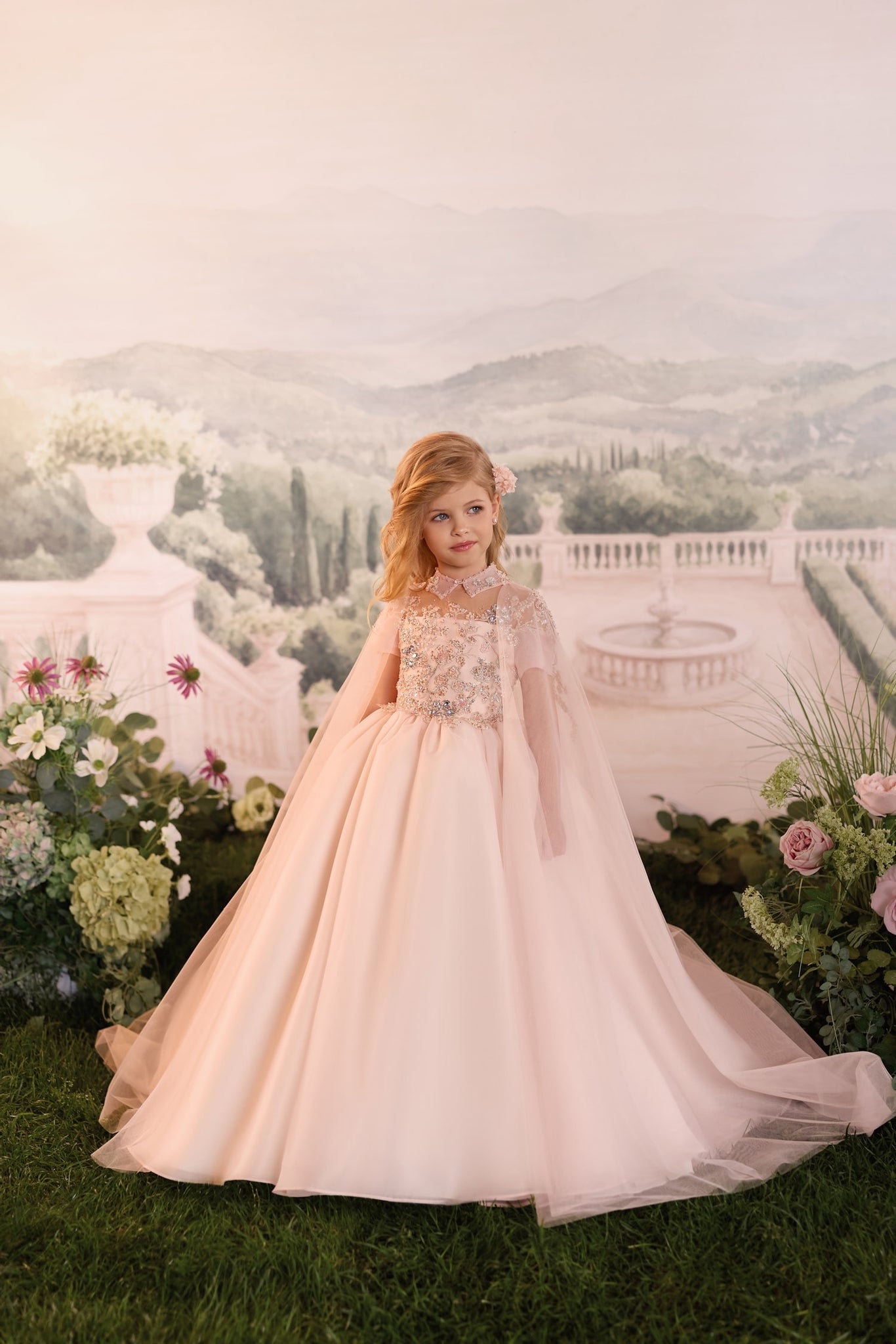 Young girl wearing a pale pink floor length ball gown with a sheer cape and floral embellishments