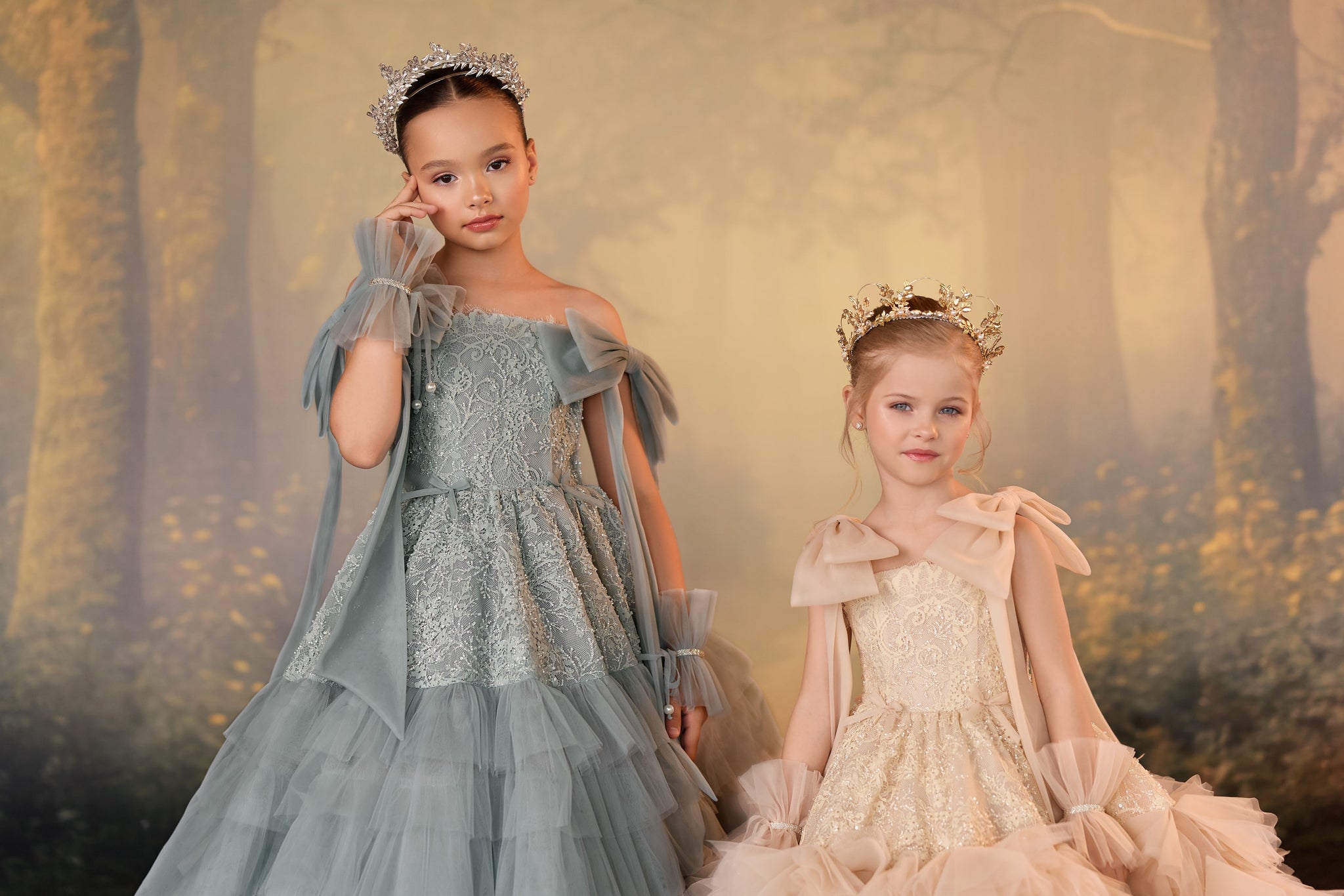 Two girls wearing formal lace dresses and tiaras against a forest background