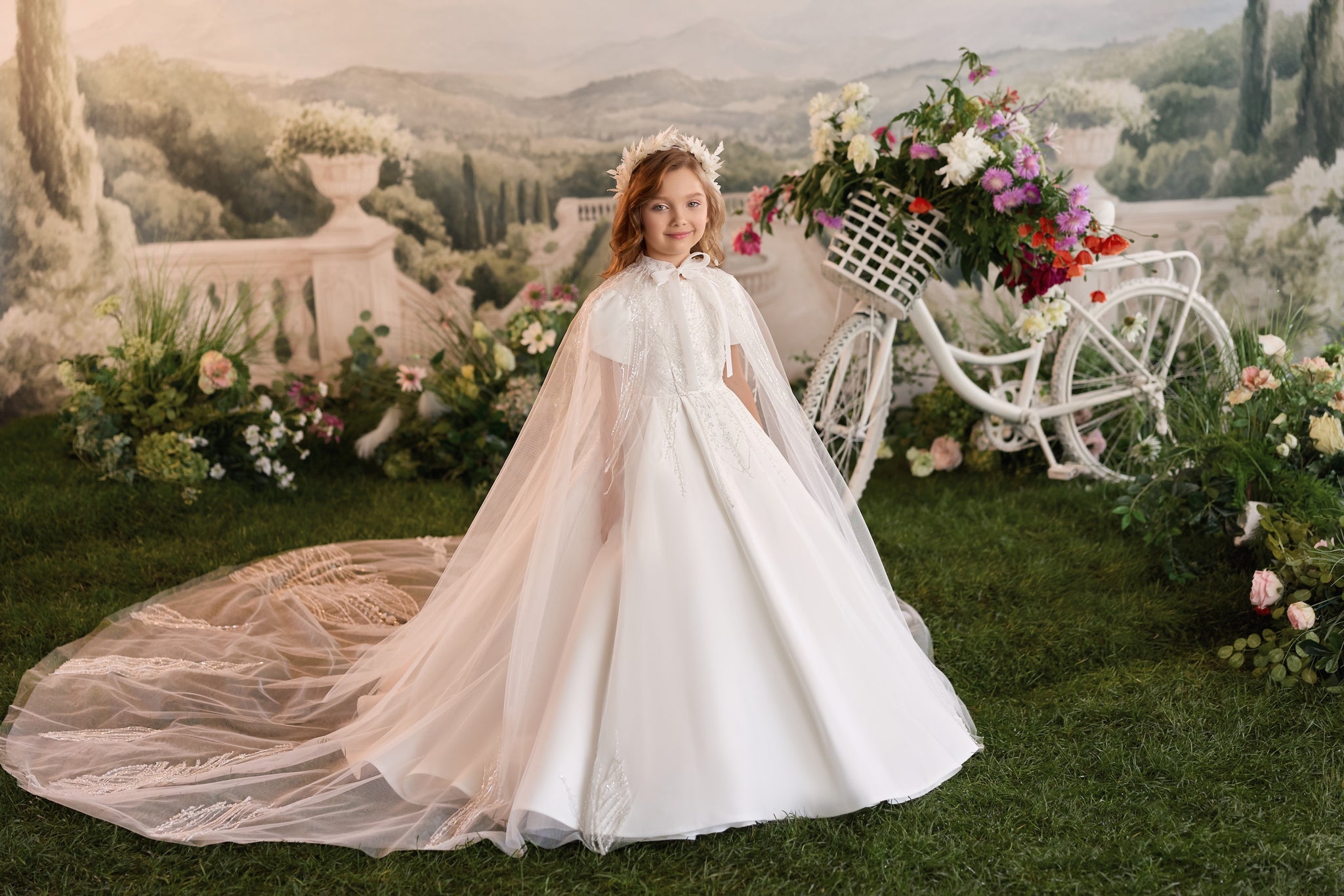 Girl wearing a white communion dress with a long cape and floral headpiece standing next to a white bicycle
