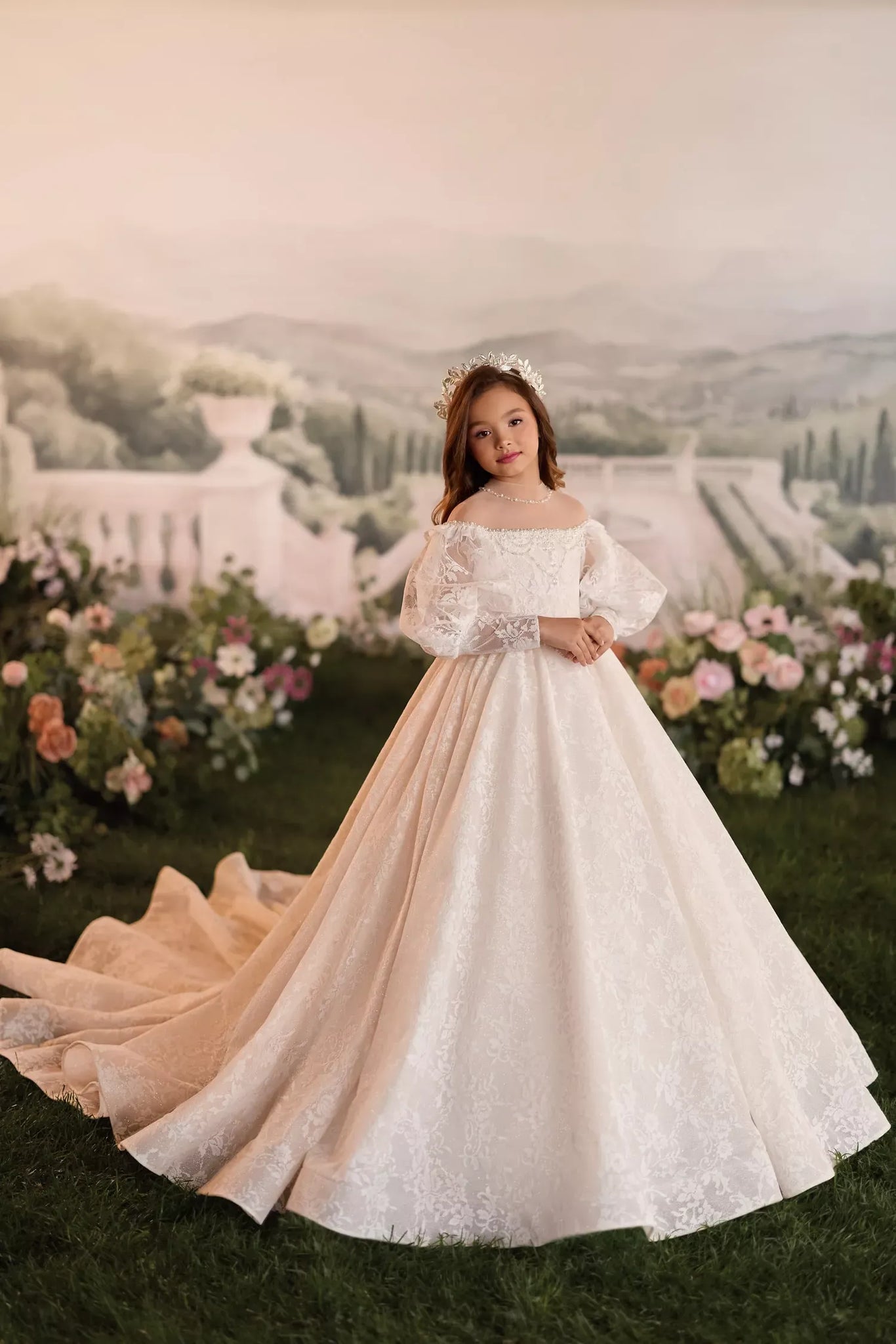 Girl wearing a white lace ball gown with puff sleeves and a tiara standing in a garden setting
