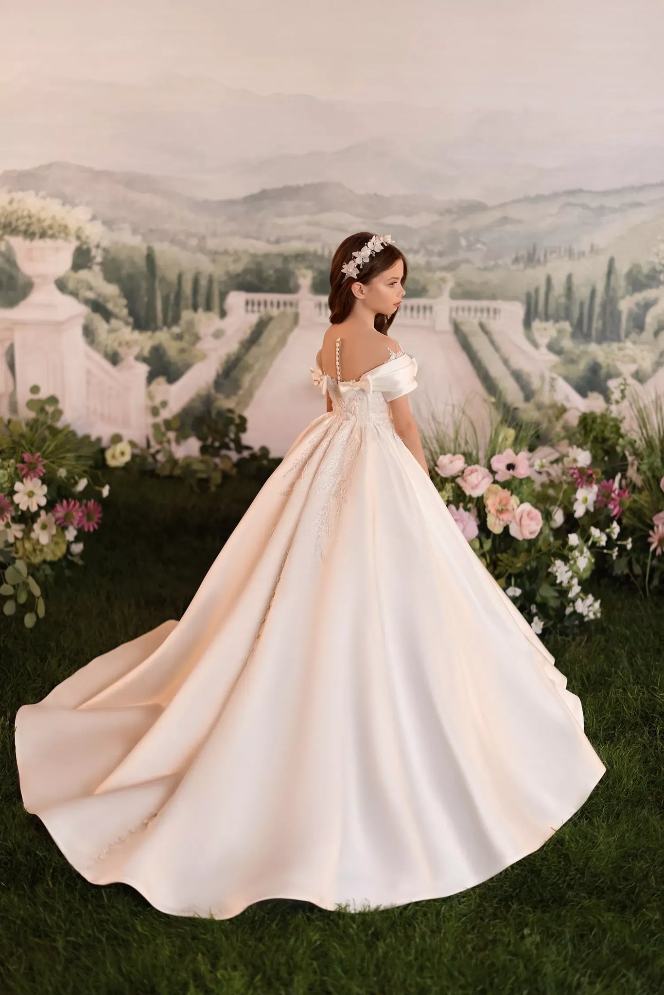 Girl wearing a white off-the-shoulder ball gown with a floral headband standing in a garden setting