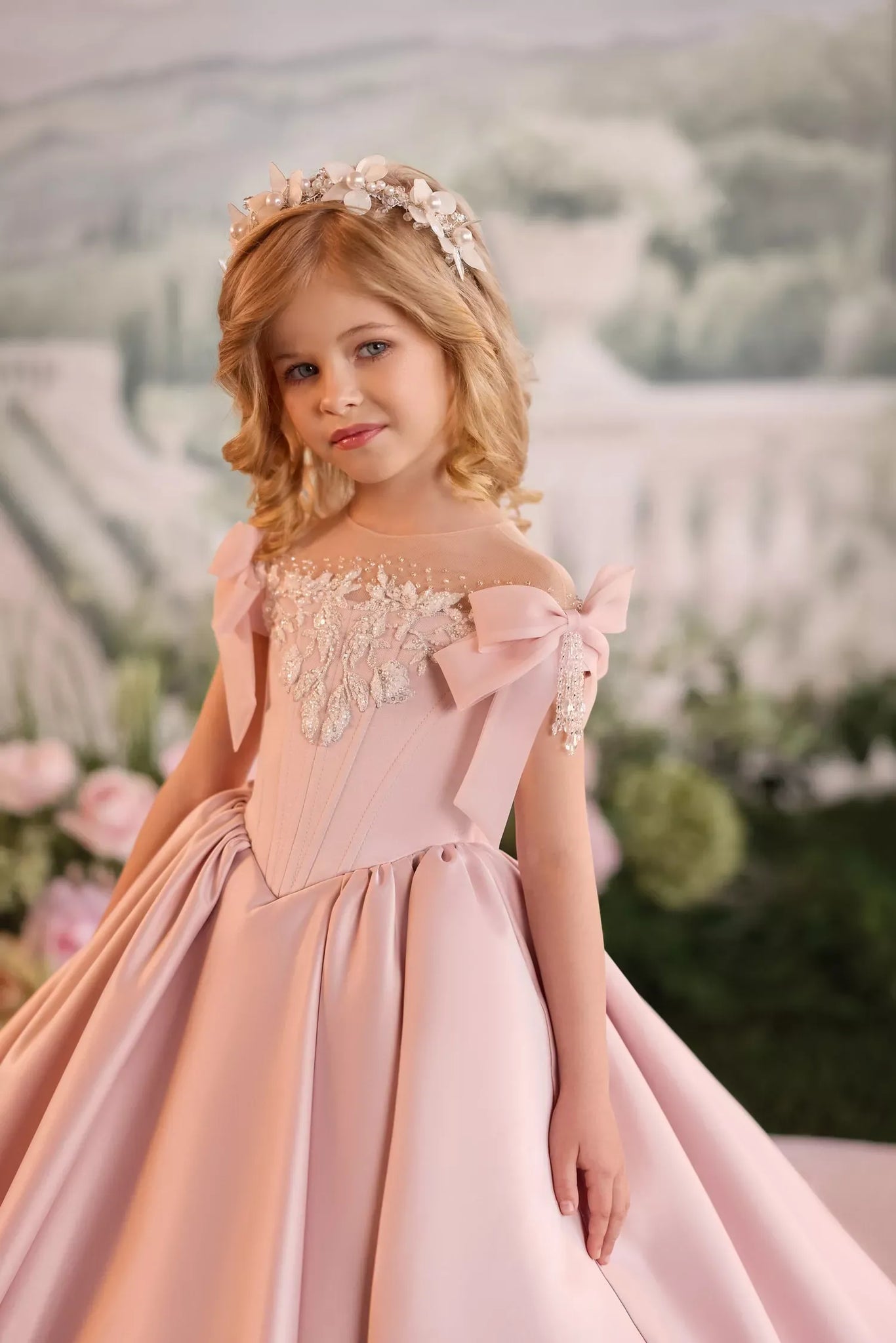 A young girl with blonde hair wearing a pink off-the-shoulder ball gown with lace detailing and a matching floral crown, posed against a soft, blurred garden backdrop.