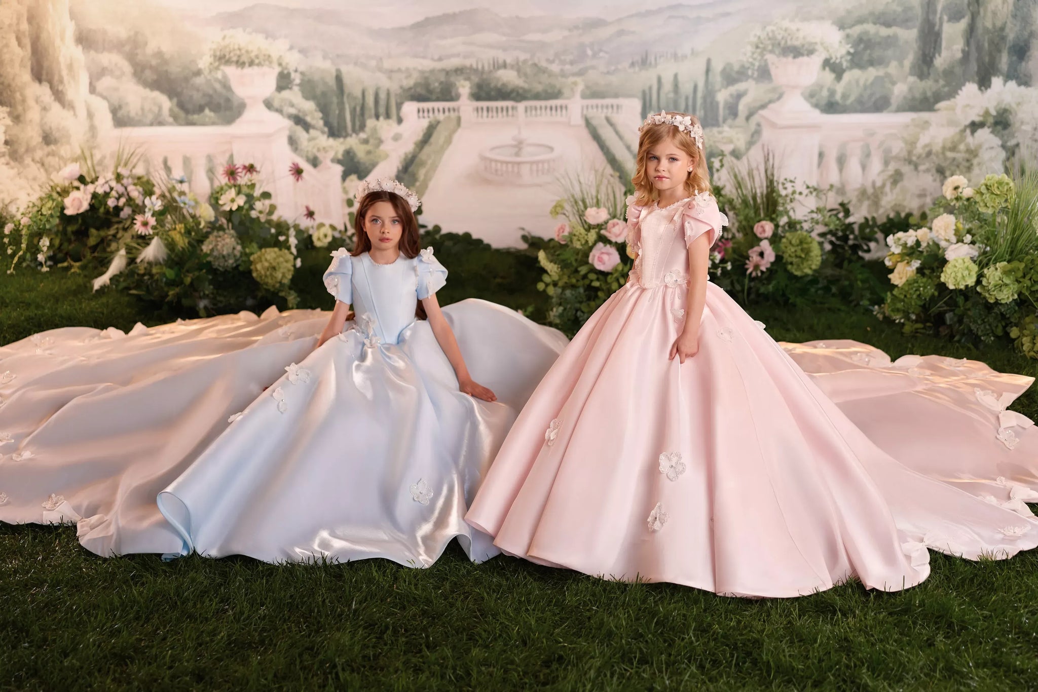 Two girls wearing long pastel blue and pink ball gowns with floral headpieces in a garden setting