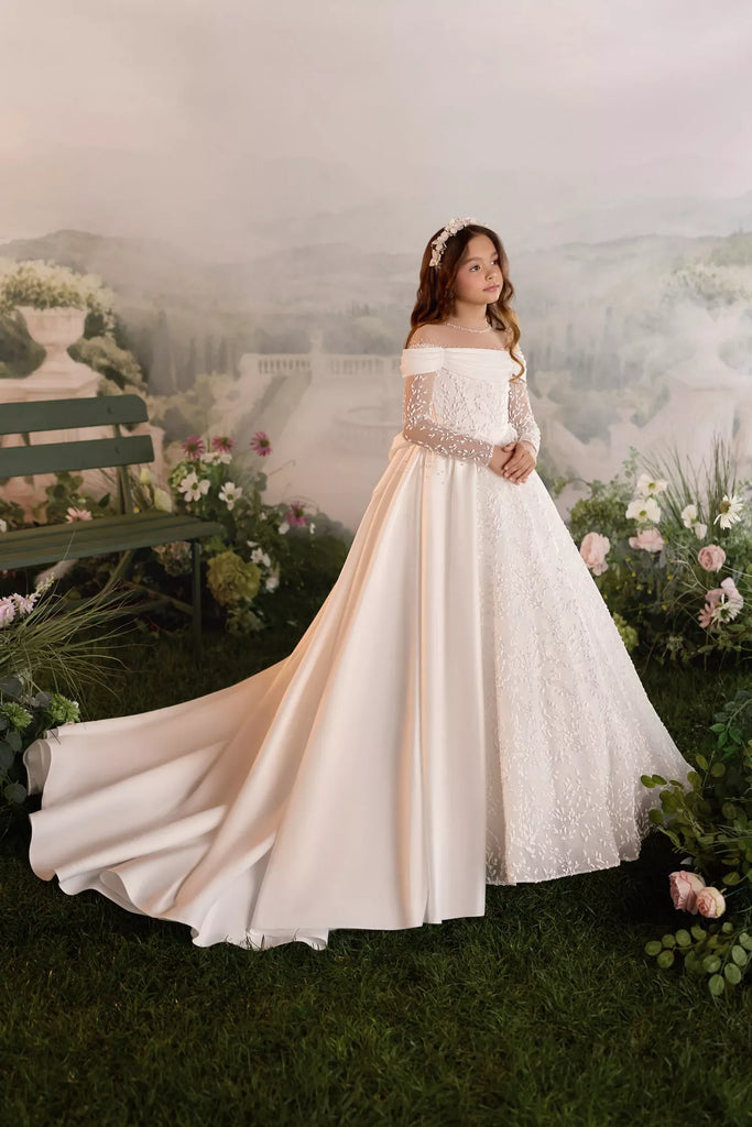 Girl wearing a white off the shoulder ball gown with lace sleeves and a long train in a garden setting | Girls Formal Dress