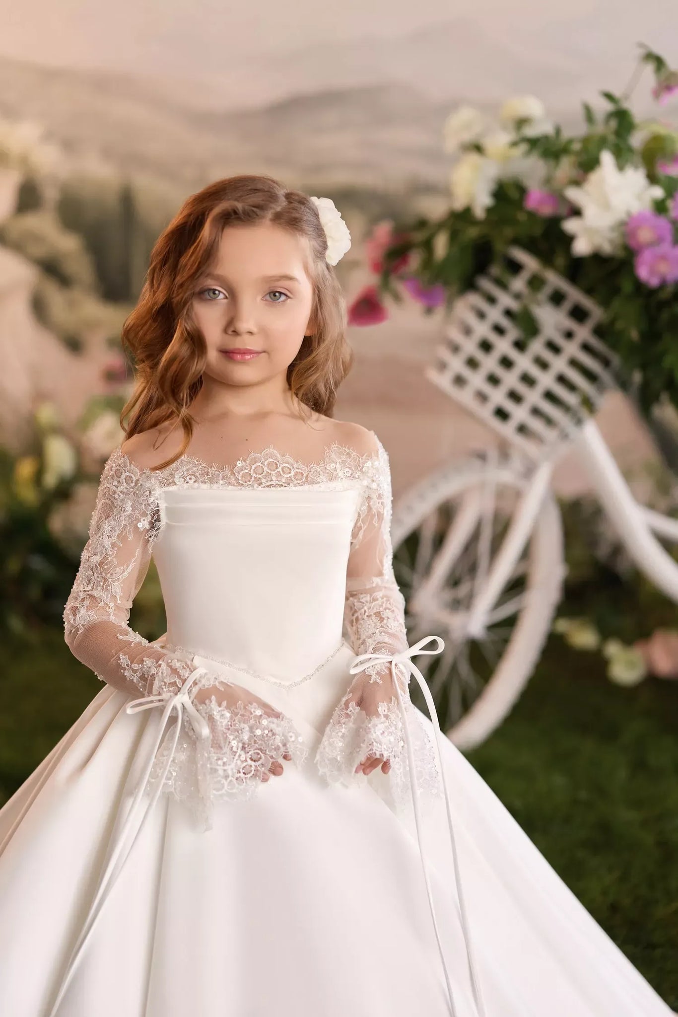 Girl wearing a white off the shoulder dress with lace sleeves and a white bicycle with flowers in the background