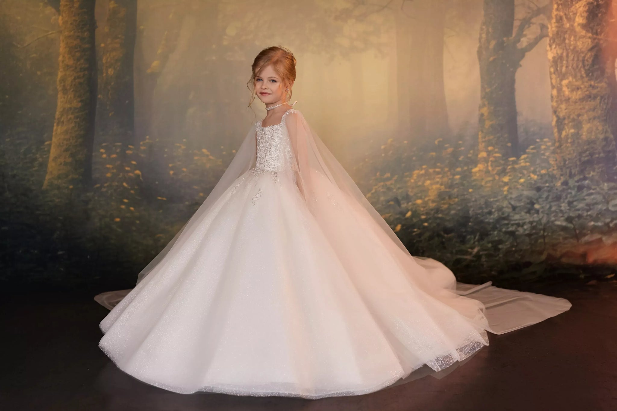 A young girl with red hair stands in a white ball gown with a sheer tulle cape, posed in front of a forest-themed backdrop.