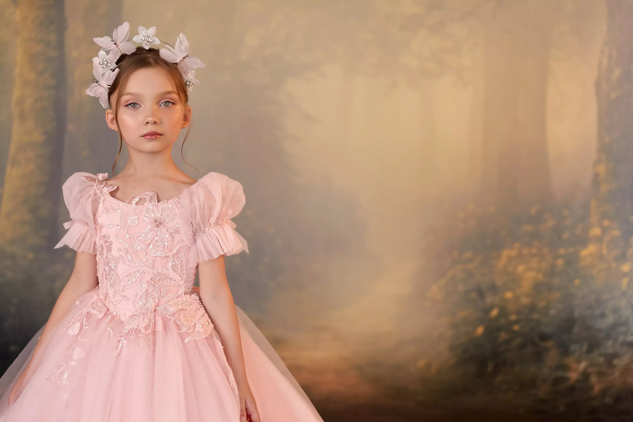 A young girl wearing a light pink fairy-tale style dress with ruffled sleeves and a matching white floral headpiece, posed against a soft, misty forest background.