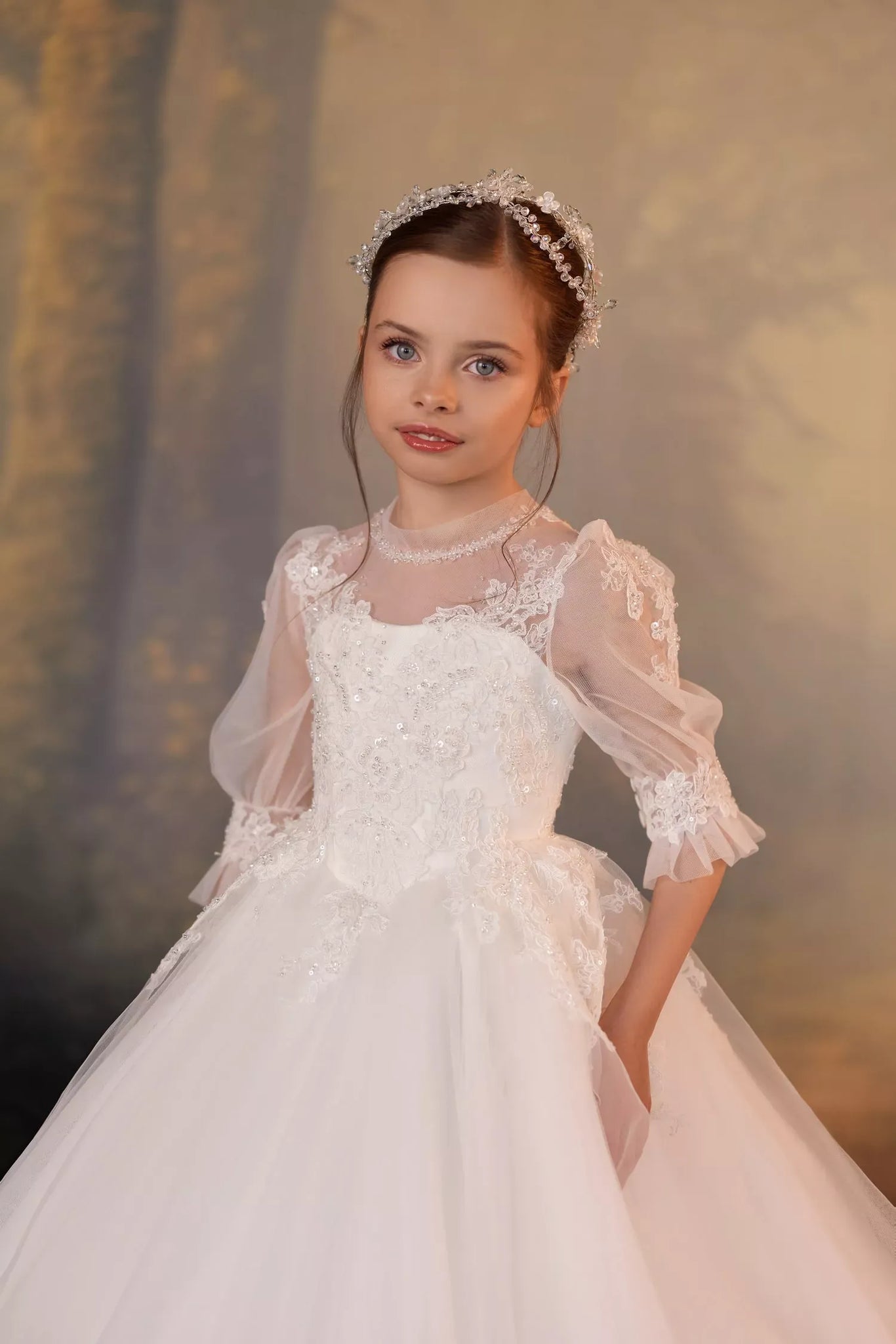 Girl wearing a lace ball gown with puff sleeves, a pearl headpiece, and a full tulle skirt | Girls Formal Dress
