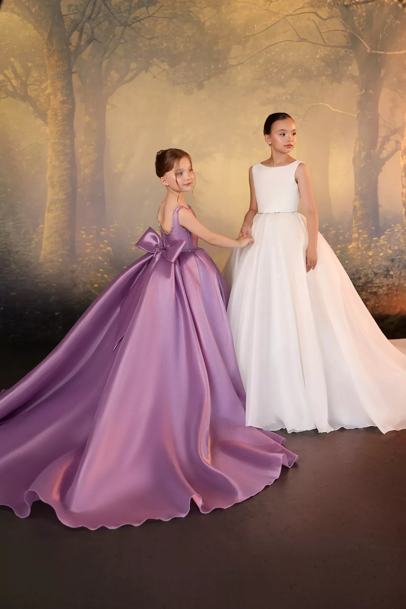 Two girls wearing formal floor length dresses in lilac and ivory standing in front of a forest backdrop | Girls Formal Dress