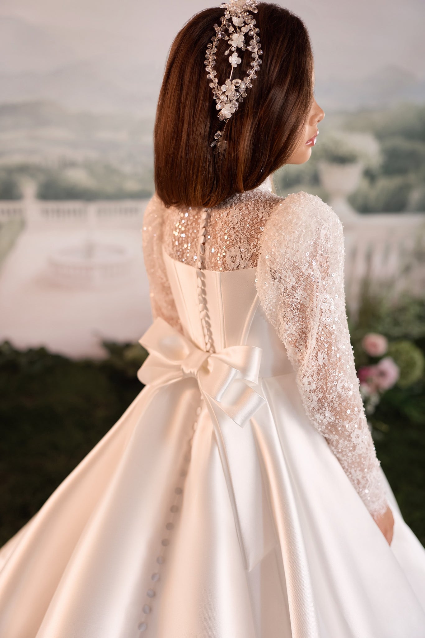 A girl shown from behind wearing a long-sleeved ivory wedding dress with a large bow at the back and a matching floral headpiece, set against a soft, blurred outdoor garden backdrop.