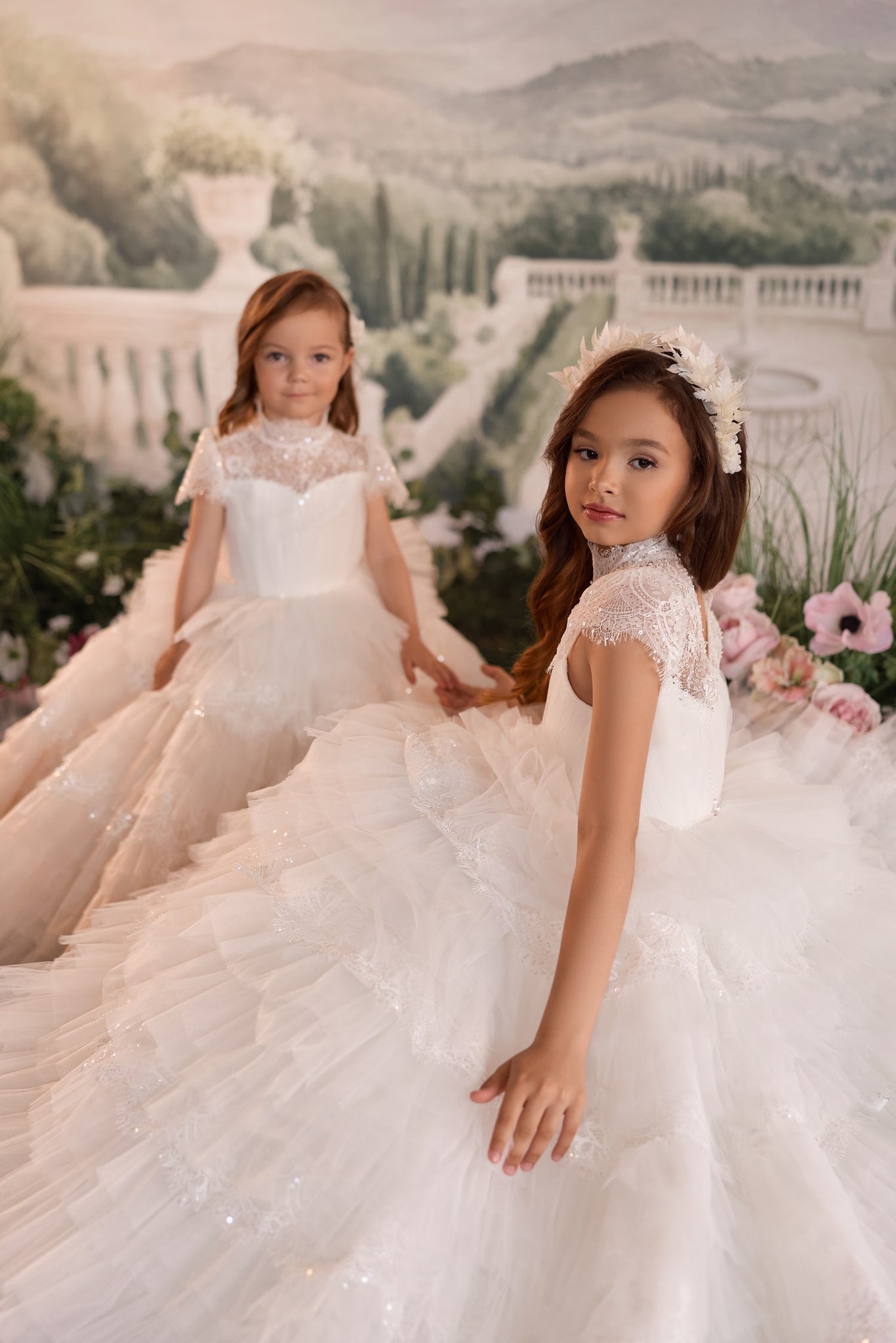 Two girls wearing white lace and tulle formal dresses with floral headpieces in a garden setting