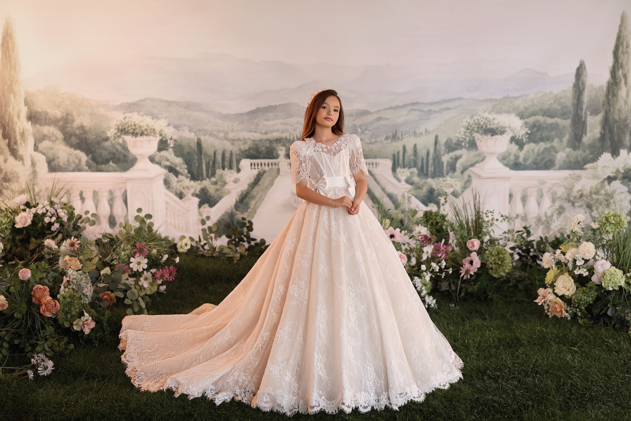 Girl wearing a white lace ball gown with a train standing in front of a garden backdrop